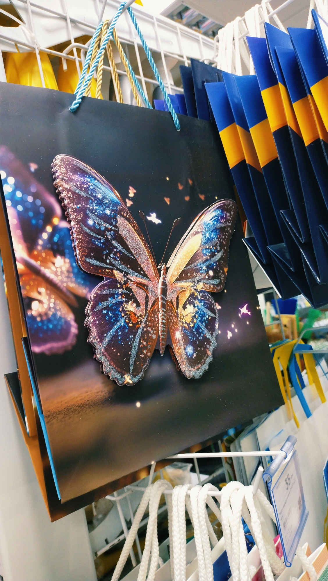 A close-up, eye-level shot captures a black gift bag featuring a large, detailed image of a butterfly with iridescent blue and gold wings. The butterfly is positioned centrally on the bag, which is hanging from a white wire rack. The bag has two twisted rope handles, one blue and one gold, attached to the top. To the right of the gift bag, a stack of blue and yellow paper bags are neatly arranged, with their pointed tops visible. In the background, more white rope handles of other gift bags hang from the rack. A price tag reading "35-" is clipped to the wire rack, partially obscured by the white rope handles. The overall environment appears to be a retail setting, possibly a store, with other merchandise visible in the background, though out of focus. The lighting is bright, and the image is slightly grainy.