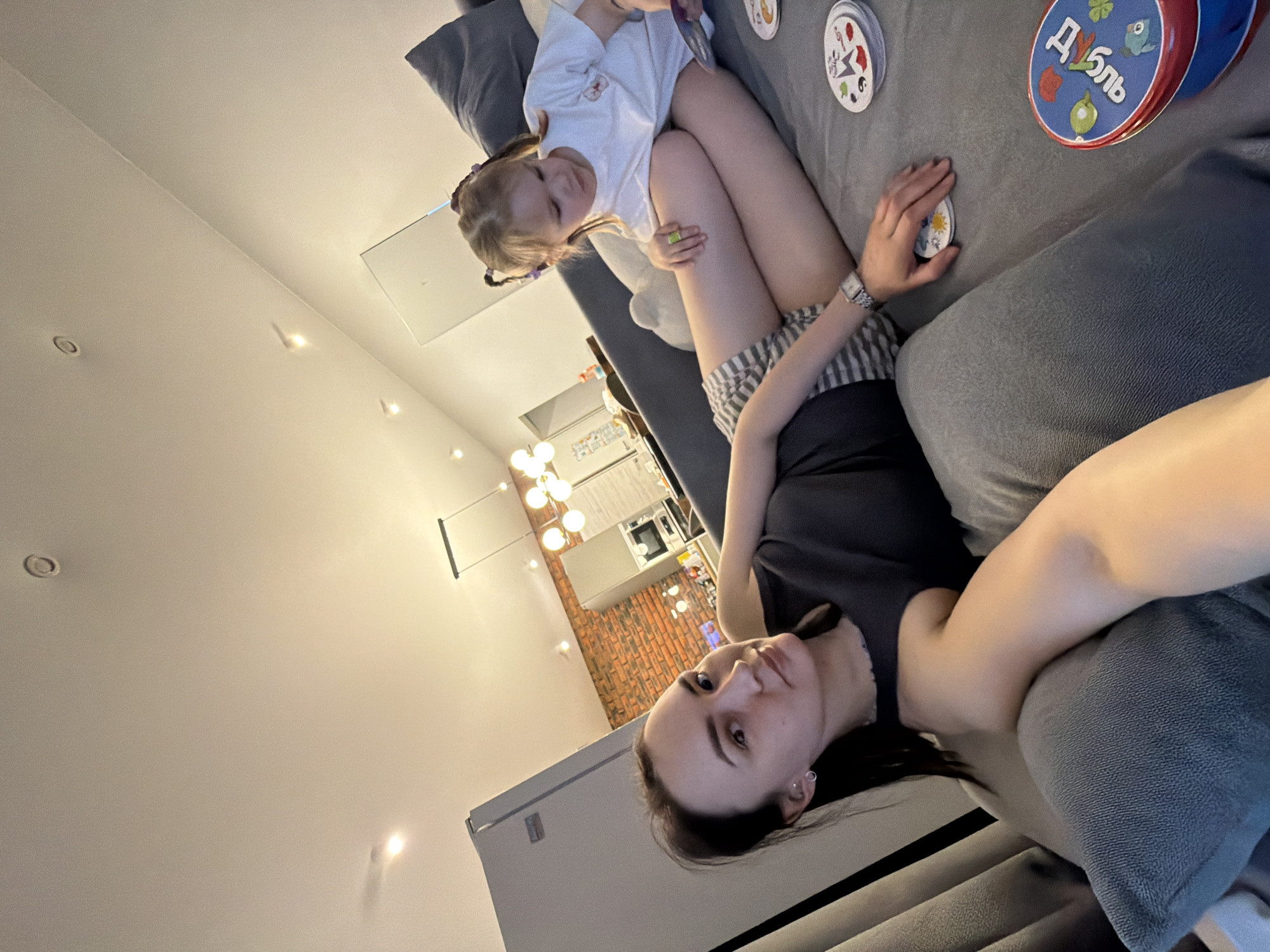 A photograph depicts a woman and a young girl on a gray sofa, seemingly engaged in a card game. The woman is positioned closer to the camera, facing towards the viewer. The young girl is situated further back on the sofa. Playing cards are scattered on the sofa's surface, and a circular object with Cyrillic script is also present. The photograph appears to have been taken indoors, with a kitchen visible in the background. The photograph was taken in Chaikovsky, Russia.