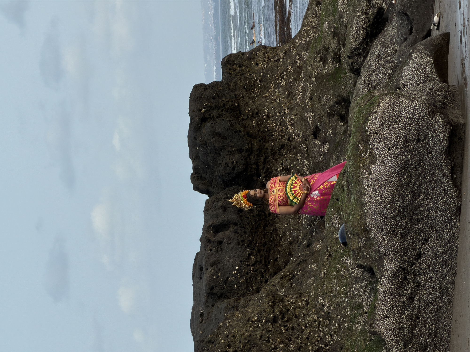 A woman in traditional Balinese attire, including a headdress and a pink and gold dress, sits on a rocky outcrop by the ocean. She holds a green and gold fan in her hands. The rocks are covered in small shells and patches of moss. In the background, the ocean has waves, and a few surfers are visible in the distance. The sky is overcast with light clouds. The location appears to be a beach in North Kuta, Indonesia.