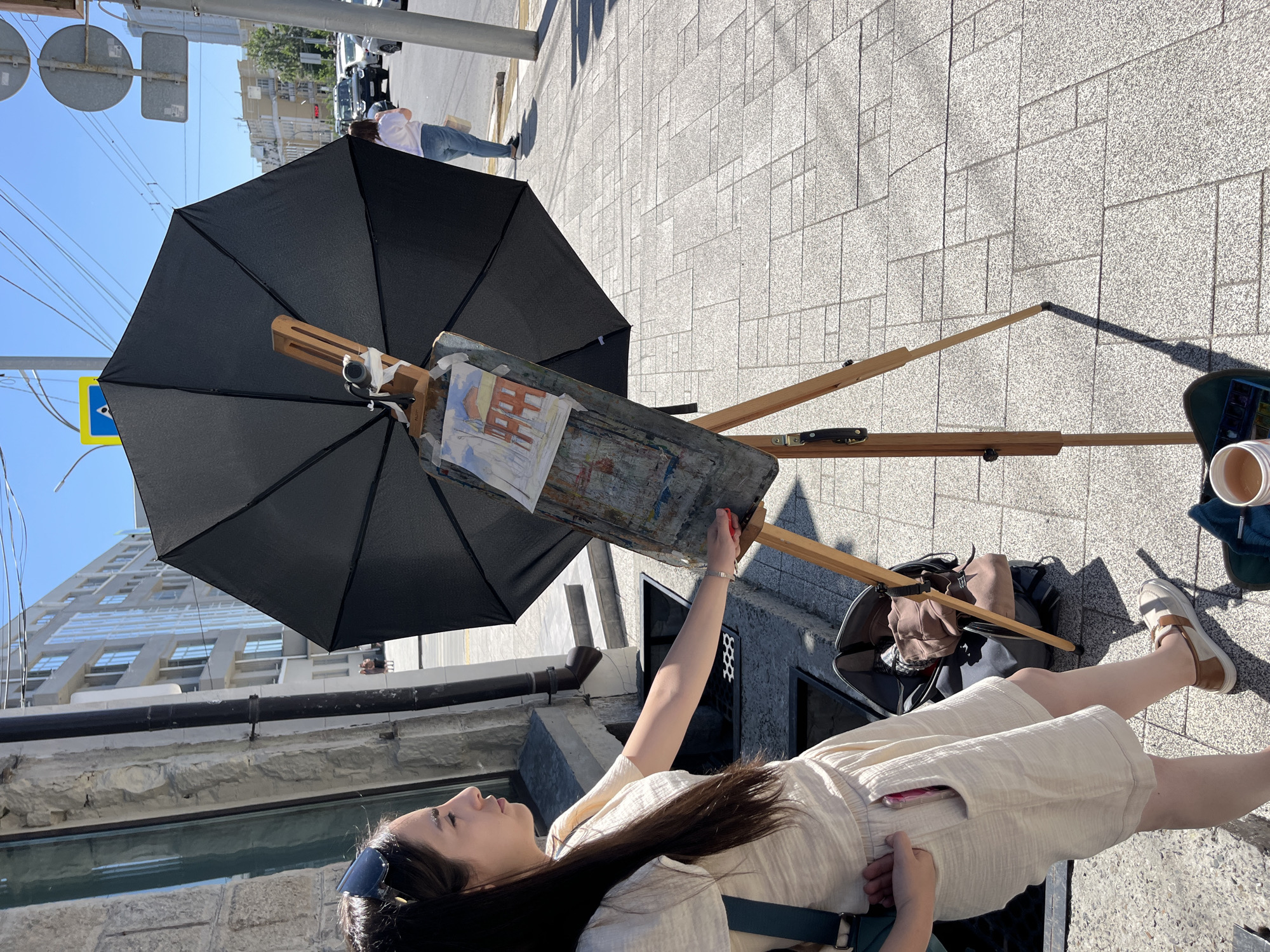 In an outdoor urban setting, a young woman with long dark hair and sunglasses on her head stands to the left of a wooden easel displaying a partially completed painting of a building. She is wearing a light-colored, loose-fitting outfit and has a cell phone in her pocket. Next to the easel, an open black umbrella is propped up, presumably to shade the artwork and artist from the sun. A bag and art supplies, including a cup of liquid and a palette, are visible on the ground near the easel. In the background, a street with moderate traffic and buildings can be seen. The ground is paved with light-colored rectangular tiles, and the sky is clear and blue.