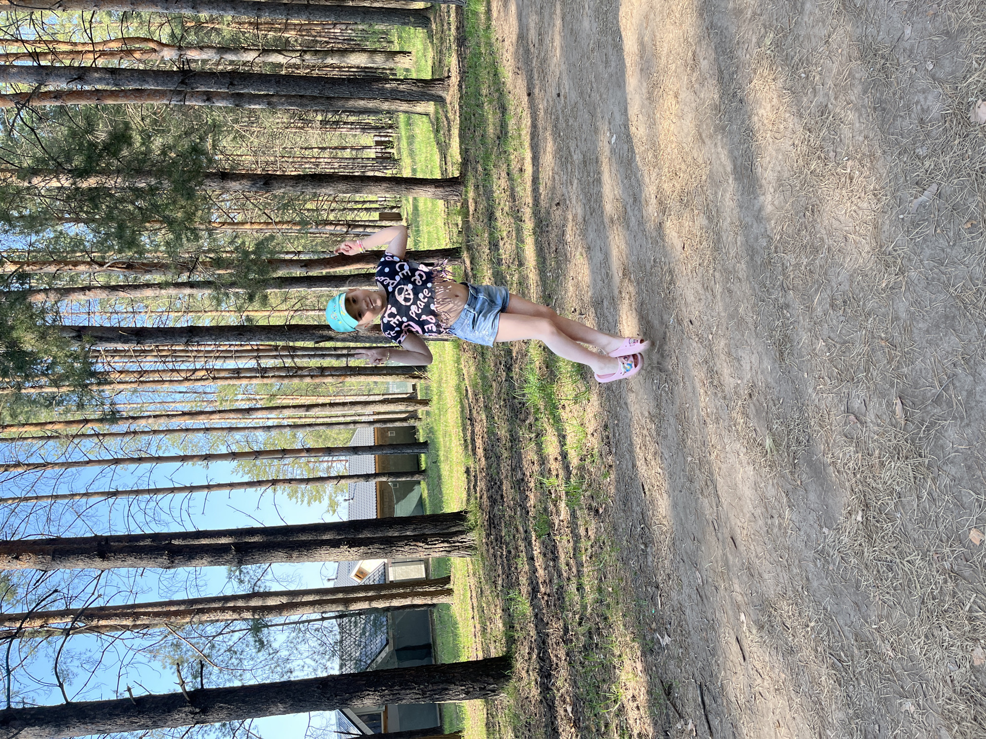 A child wearing a blue cap, a black shirt with white writing, blue shorts, and pink sandals is lying on a dirt path in a forest in Chaikovsky, Russia. The path is surrounded by tall pine trees. A small building is visible between the trees.