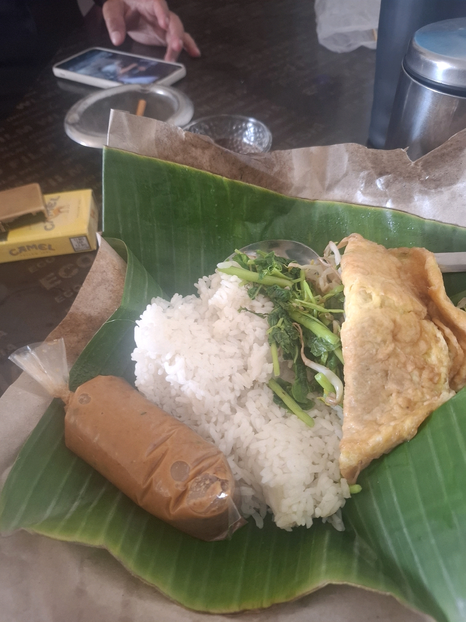 In Ciheulang Tonggoh, Indonesia, a close-up shot captures a simple meal served on banana leaves. The meal consists of white rice, a piece of folded omelet, and some cooked vegetables, possibly water spinach and bean sprouts. A plastic packet contains a portion of peanut sauce. To the left, a portion of a cigarette pack, identified by the "Camel" logo, is visible. In the background, out of focus, a hand is reaching towards a smartphone placed on a table next to an ashtray containing a cigarette butt. The setting appears to be a casual dining environment, likely outdoors or in a semi-open area, given the presence of a metallic container and a paper wrap.