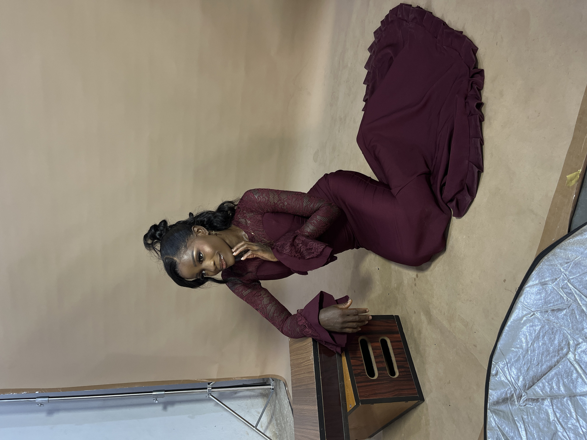 In Gura Top, Nigeria, a young Black woman is posing for a photo shoot. She is kneeling on the floor in a studio setting, wearing a long, deep red gown with lace sleeves. The dress has a fitted bodice and a flowing skirt that trails behind her. Her hair is pulled up in a high ponytail, with some strands framing her face. She is looking to her left, with one hand resting on her chin and the other on a wooden prop next to her. The prop is a rectangular wooden box with two horizontal openings on the side. The background is a plain, light brown backdrop. There is also a silver reflector on the floor in the lower left corner of the image. The lighting is soft and even, suggesting a professional studio setup.