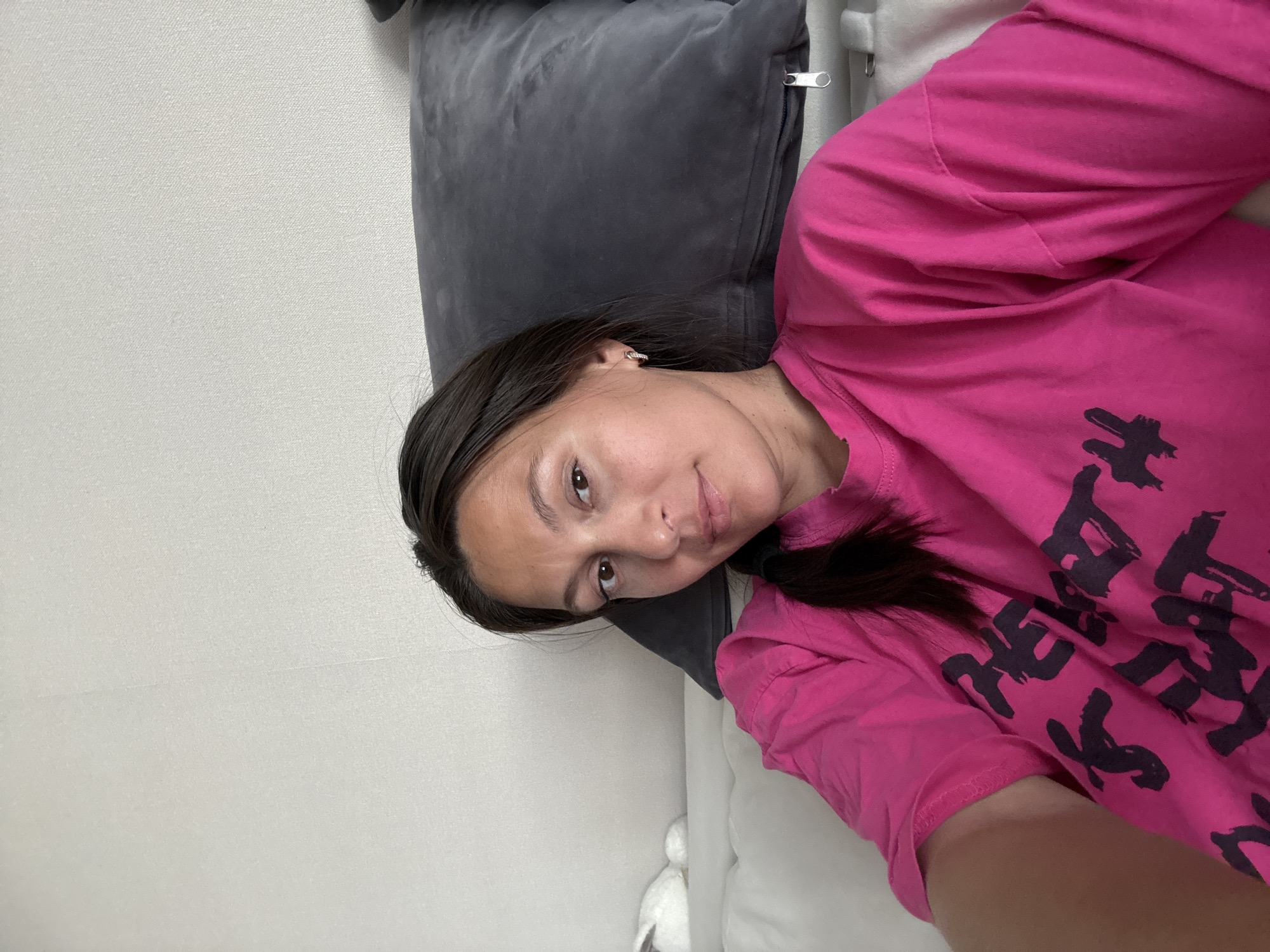 A woman lies on a bed. She is wearing a pink shirt with black lettering. She is resting her head on a grey pillow. The background consists of a plain white wall. A stuffed animal sits on the bed nearby. The exact location in Chaikovsky, Russia, cannot be determined from the image.