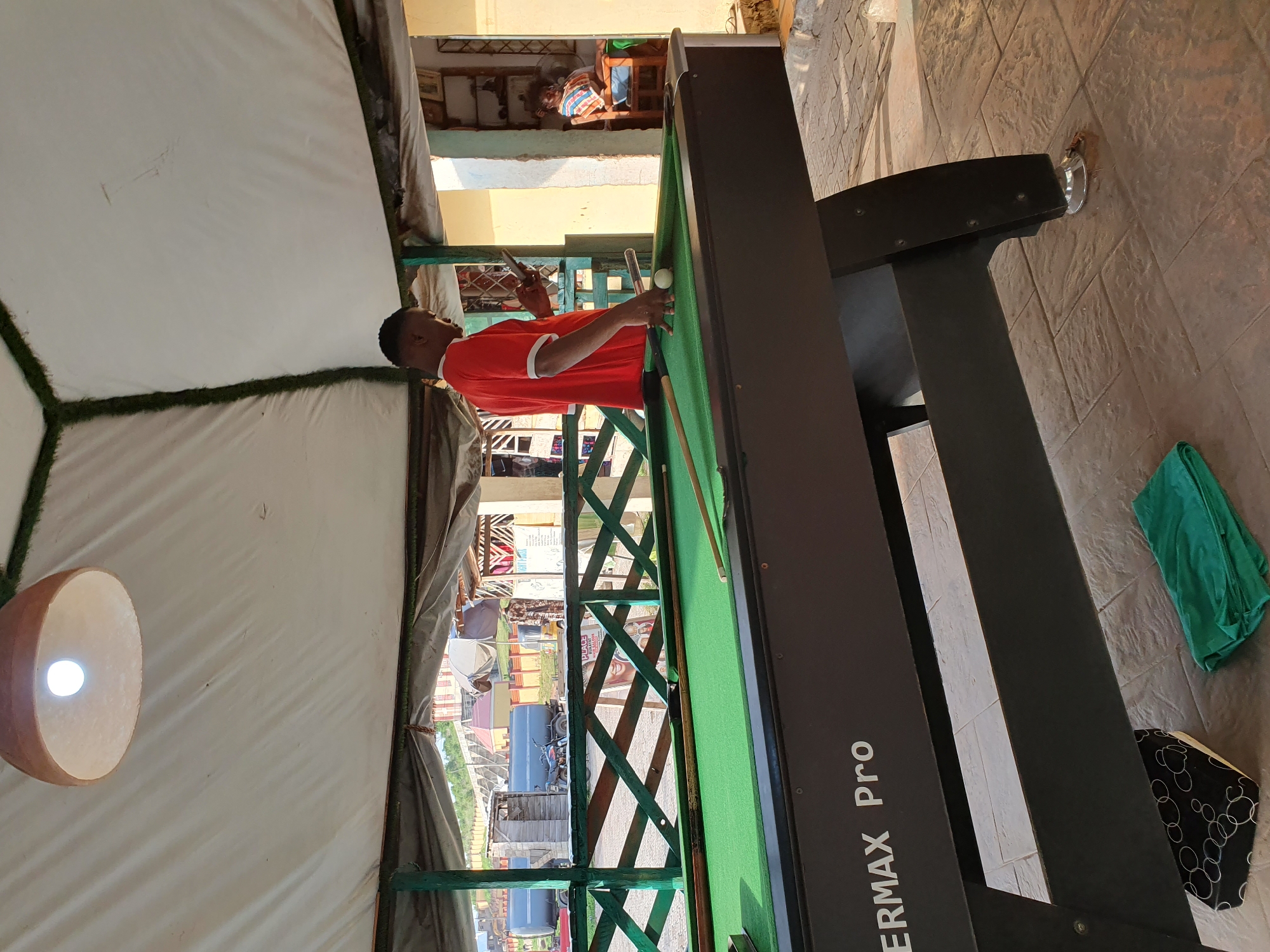 In Lokoja, Nigeria, a man in a red shirt stands by a billiard table, holding a phone. The table, labeled "ERMAX Pro," has a green felt surface and is set up outdoors, possibly under a canopy. A billiard cue rests on the table near a white ball. In the background, a woman is seated in a chair, and beyond her, buildings and a road are visible. The environment appears to be a recreational area, with a paved ground and a wooden fence.