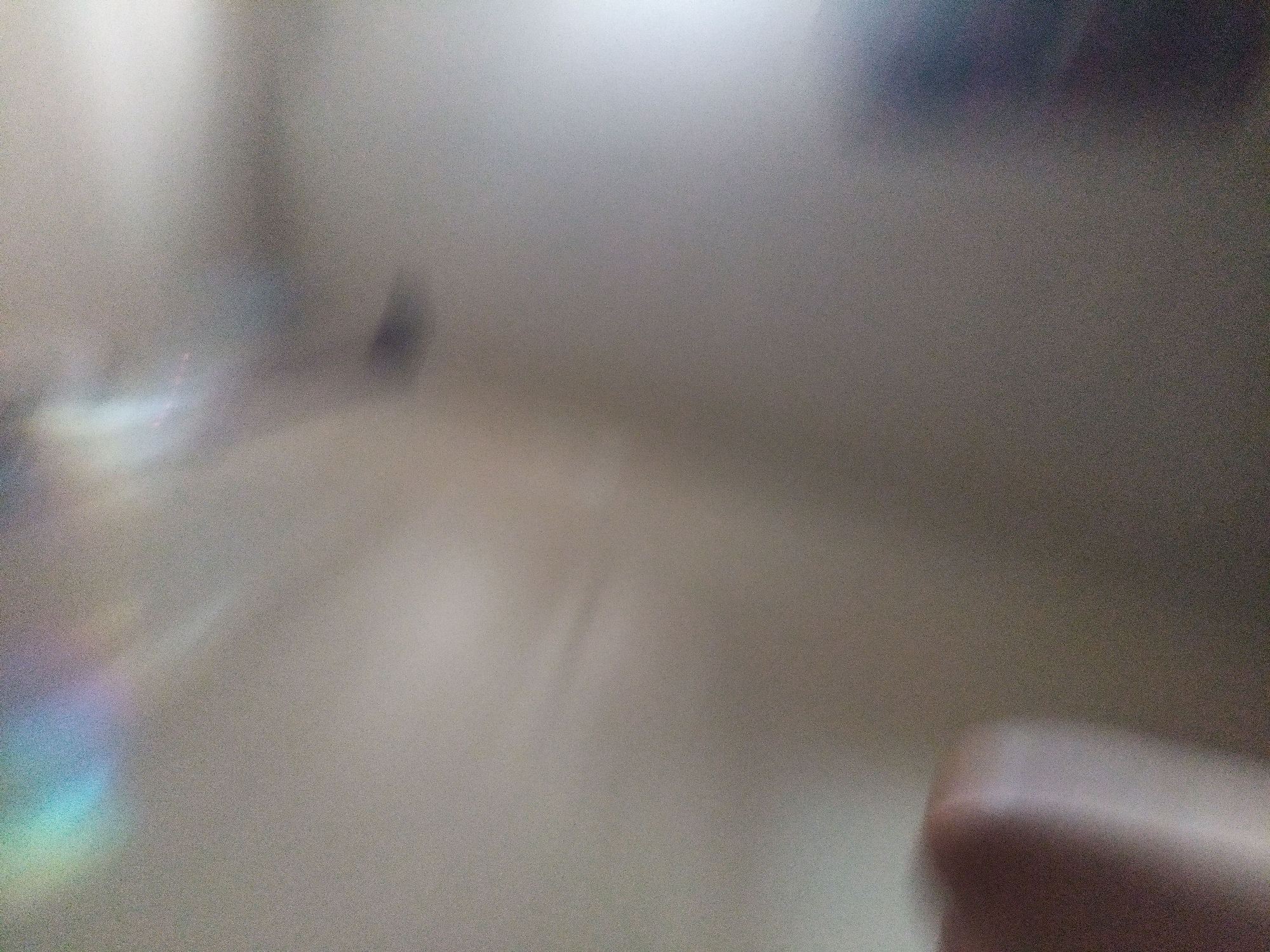 The image is a blurred, grainy photograph with a shallow depth of field. A rainbow-like prism effect is visible in the upper left quadrant. A blurred, pale, elongated object, possibly a hand or arm, occupies the lower left portion of the frame. The background is an indistinct, neutral tone, suggesting an indoor environment. There are no discernible landmarks or features that would place this image in Kragilan, Indonesia.