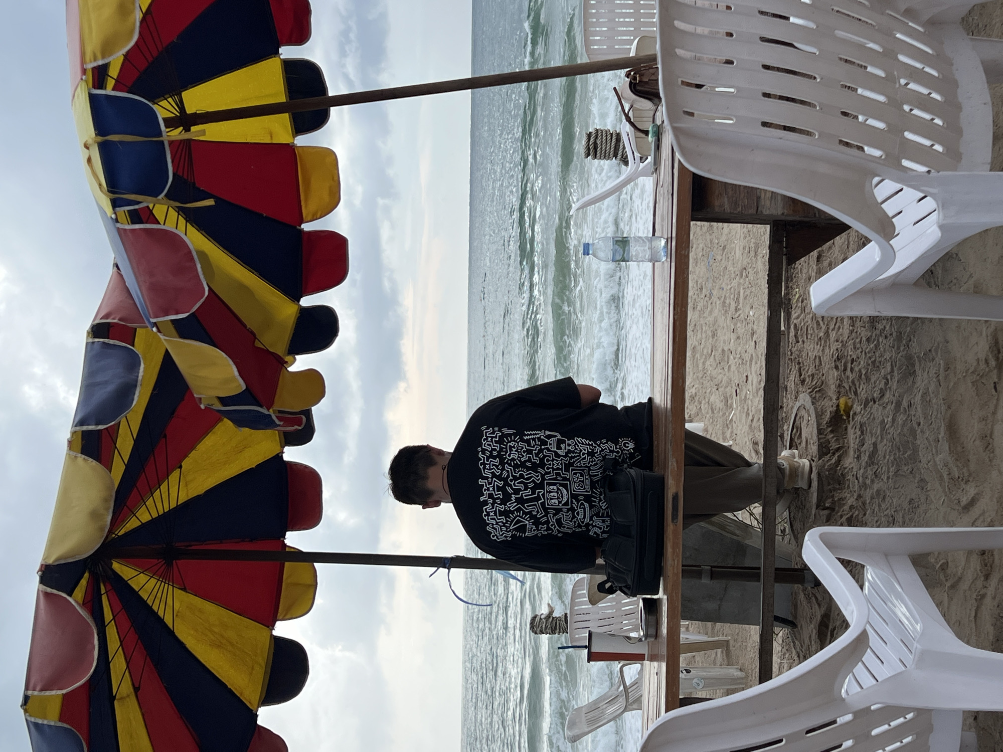 A person in a black t-shirt with white designs is seated at a wooden table on a beach. The beach is at the edge of the ocean with a cloudy sky above. A colorful beach umbrella is overhead, and white plastic chairs are around the table. A bottle of water sits on the table next to a black bag. The location is unspecified.