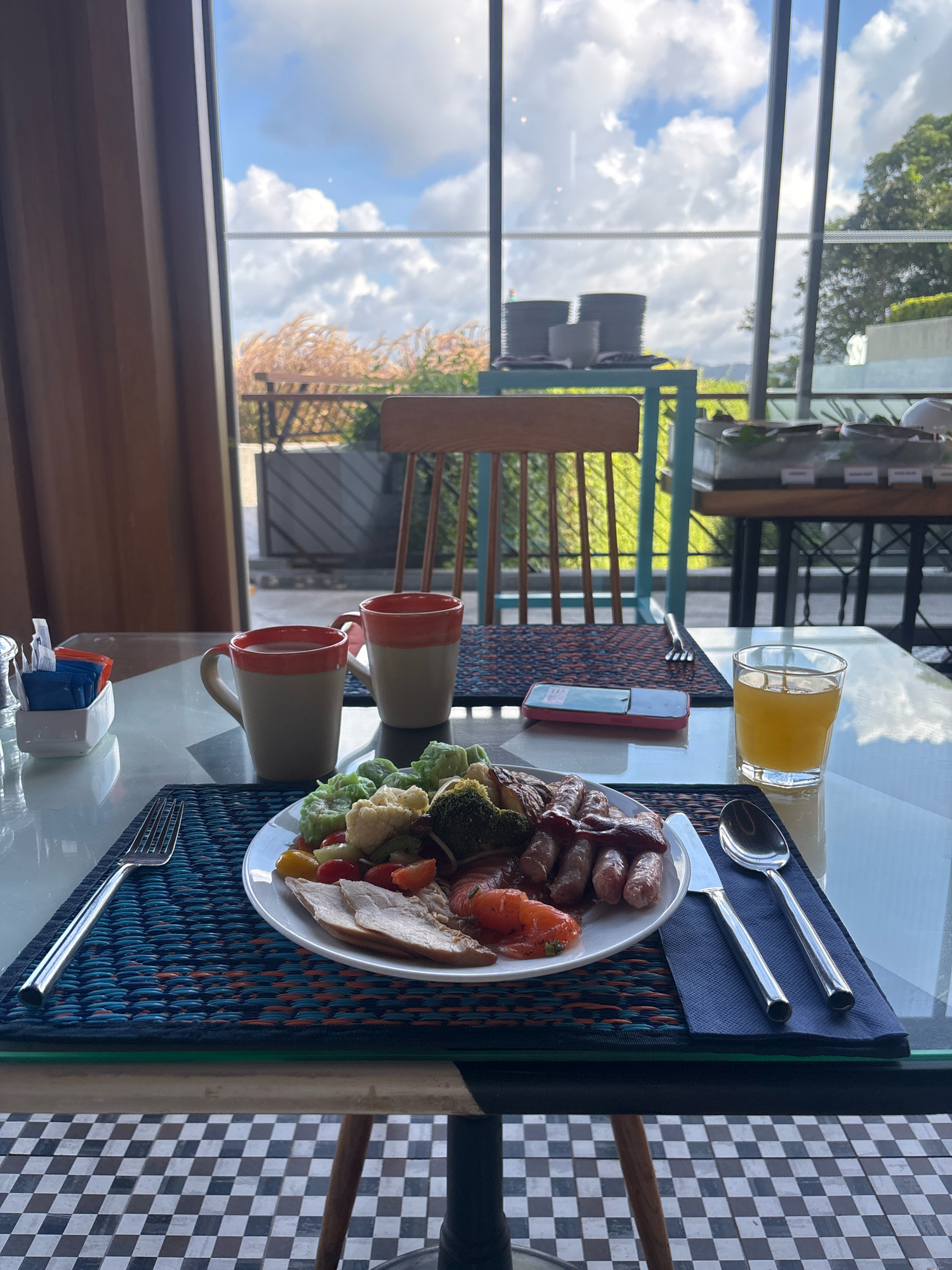 An overhead, close-up shot shows a table laid out for breakfast in a hotel buffet setting in Karon, Thailand. The table features a plate of food, two mugs of tea or coffee, a glass of orange juice, and cutlery. The plate contains sausages, sliced turkey, roasted tomatoes, broccoli, cauliflower, and cherry tomatoes. Two mugs, one with a pink rim and the other with a reddish-orange rim, are positioned behind the plate. A glass of orange juice sits to the right of the plate. The table is set with a blue and orange placemat and a blue napkin holding a knife and spoon. A salt and pepper shaker set and sugar packets are visible on the left. In the background, through large windows, a bright, cloudy sky and lush green trees are visible, suggesting an outdoor or semi-outdoor dining area. Chairs are also visible in the background, indicating a dining establishment. The floor beneath the table is a black and white checkered pattern.
