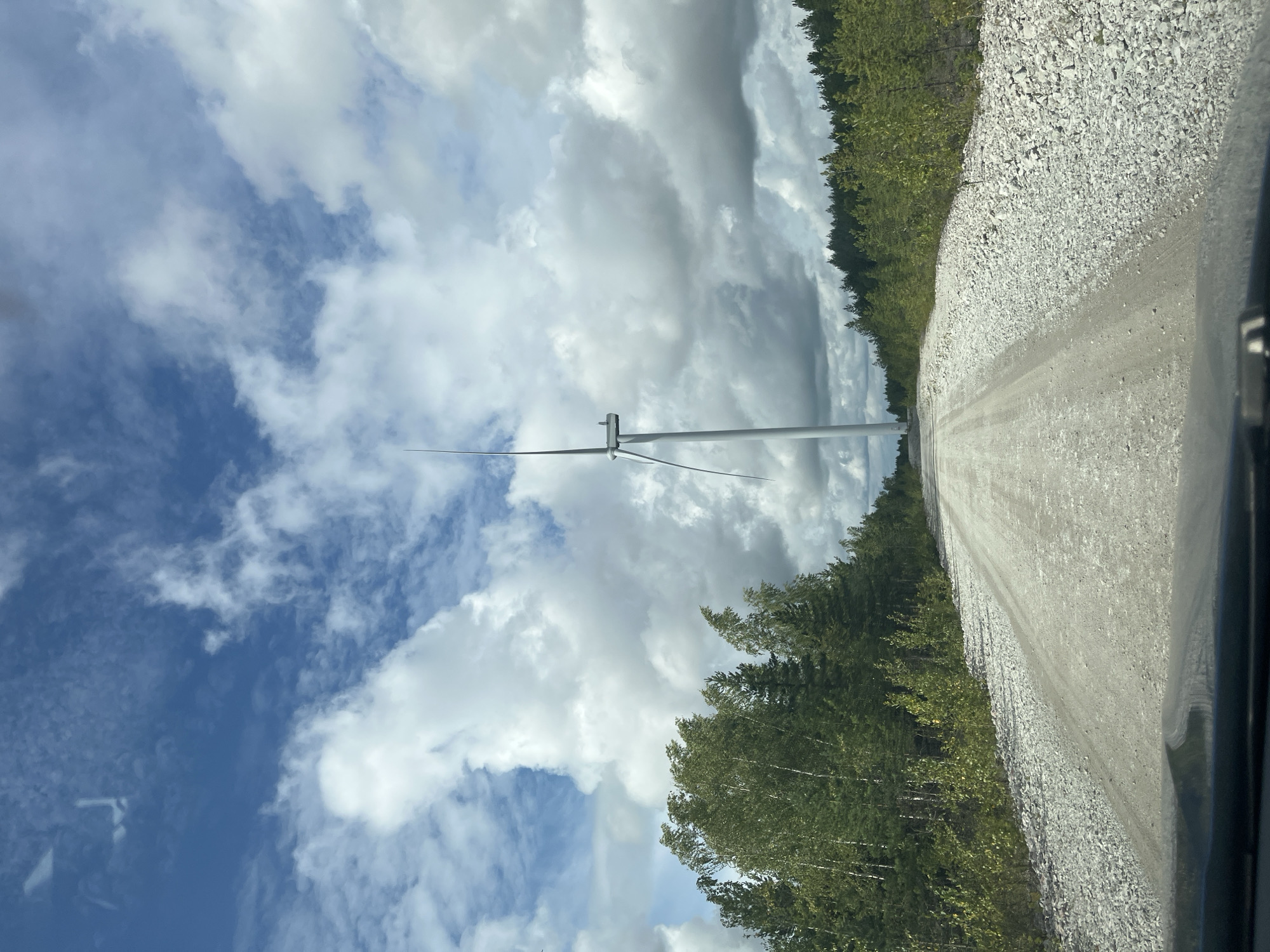 A wide shot captures a gravel road leading towards a solitary wind turbine, set against a backdrop of a bright blue sky filled with voluminous, puffy white clouds. The road, covered in light-colored gravel and dust, is flanked on either side by dense green trees. The foliage is a mix of deciduous trees with lighter green leaves, likely birch, and darker, coniferous trees. The wind turbine, a tall, slender white structure with three long blades, stands prominently in the distance, its blades positioned vertically. The scene appears to be in a rural or industrial area, possibly a wind farm, given the presence of the turbine. The overall impression is one of a serene, if somewhat stark, landscape. The location is in Aidu, Estonia, a region known for its former oil shale quarries which are now being redeveloped for renewable energy projects, including wind farms.