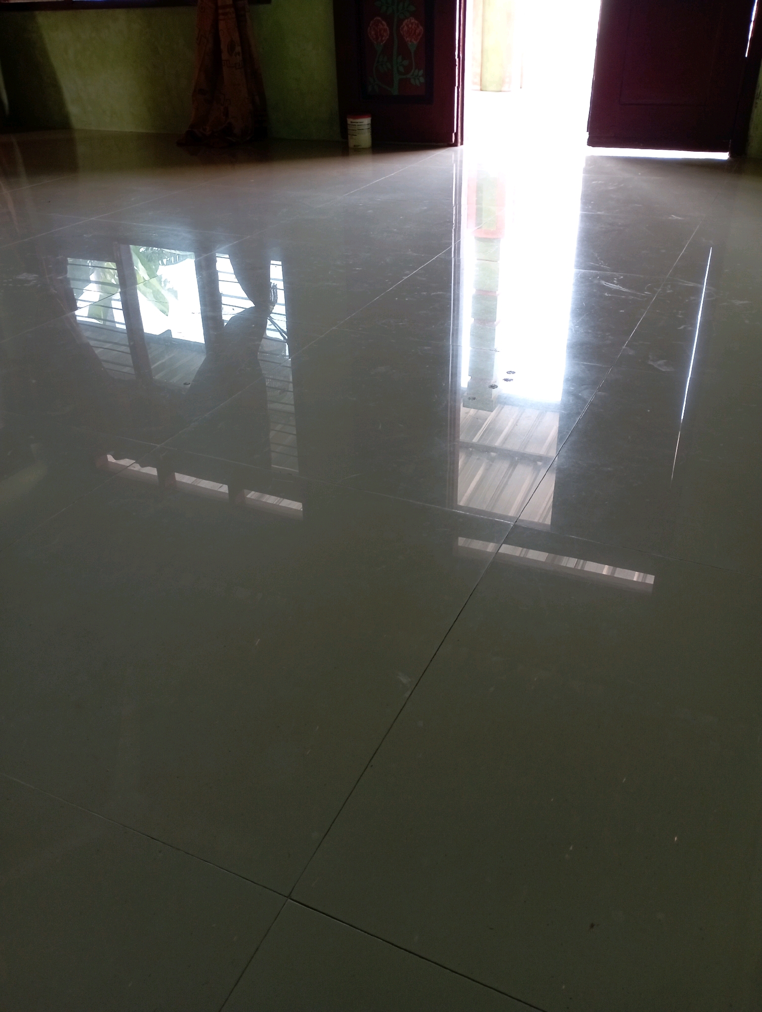 The image shows a close-up view of a polished tiled floor in what appears to be an indoor space. The floor is a dark, glossy gray with visible grout lines, creating a grid pattern. The surface reflects light and objects from above, indicating a well-lit environment.
In the upper left portion of the image, a green wall is visible, partially obscured by a dark brown curtain or fabric hanging from it. The curtain has a patterned texture. There is also a white cylindrical object, possibly a can, placed near the floor on the right side of this area.
The right side of the image shows an open doorway, leading to an intensely bright area. The light from this opening is so strong that it washes out details, but it appears to be a hallway or another room. The doorway is framed by dark red or brown door jambs.
The floor's reflection reveals elements of the environment above. In the left half of the floor, reflections show what looks like a window with horizontal blinds and greenery outside. A shadowy figure, possibly a person, is also reflected on the floor, appearing to be standing or moving near the window. The right half of the floor reflects the bright light from the doorway, along with faint outlines of what could be ceiling elements or structures.
The overall impression is one of a modern interior with a sleek, reflective floor. The strong contrast between the dark floor and the bright light from the doorway creates a dramatic effect. The scene is devoid of any explicit indications of the city of Sonobekel, Indonesia, other than the provided context.