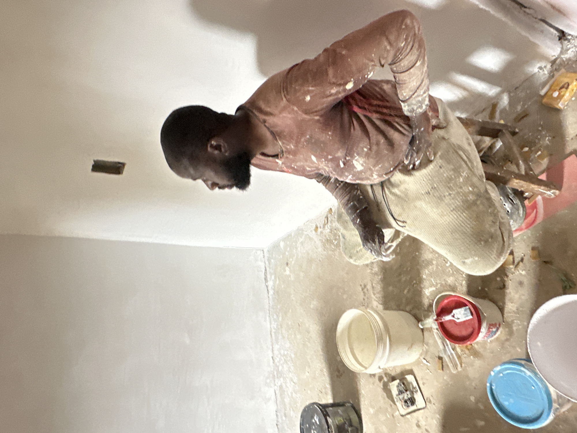 The image depicts a man engaged in renovation or construction work inside a room in Bukuru, Nigeria. The man, with dark skin and a beard, is wearing a long-sleeved, reddish-brown shirt heavily splattered with white paint, and light-colored, paint-stained trousers. He is positioned leaning forward, looking down, suggesting focus on his task.
The scene shows an indoor space with freshly plastered or painted walls and a ceiling in a light, off-white tone. A rectangular cut-out or vent is visible on the ceiling. The concrete floor is rough, dirty, and covered with paint splatters, dust, and construction debris, including wood scraps, plastic containers, and tools.
Several paint buckets or containers are scattered on the floor; one has a red lid with a paintbrush resting inside, another is white, and a third has a blue lid. The overall environment suggests an active worksite. No specific time of day or weather can be discerned, but the room is adequately lit. No legible text is visible on any objects. The man's expression appears to be one of concentration on his work.