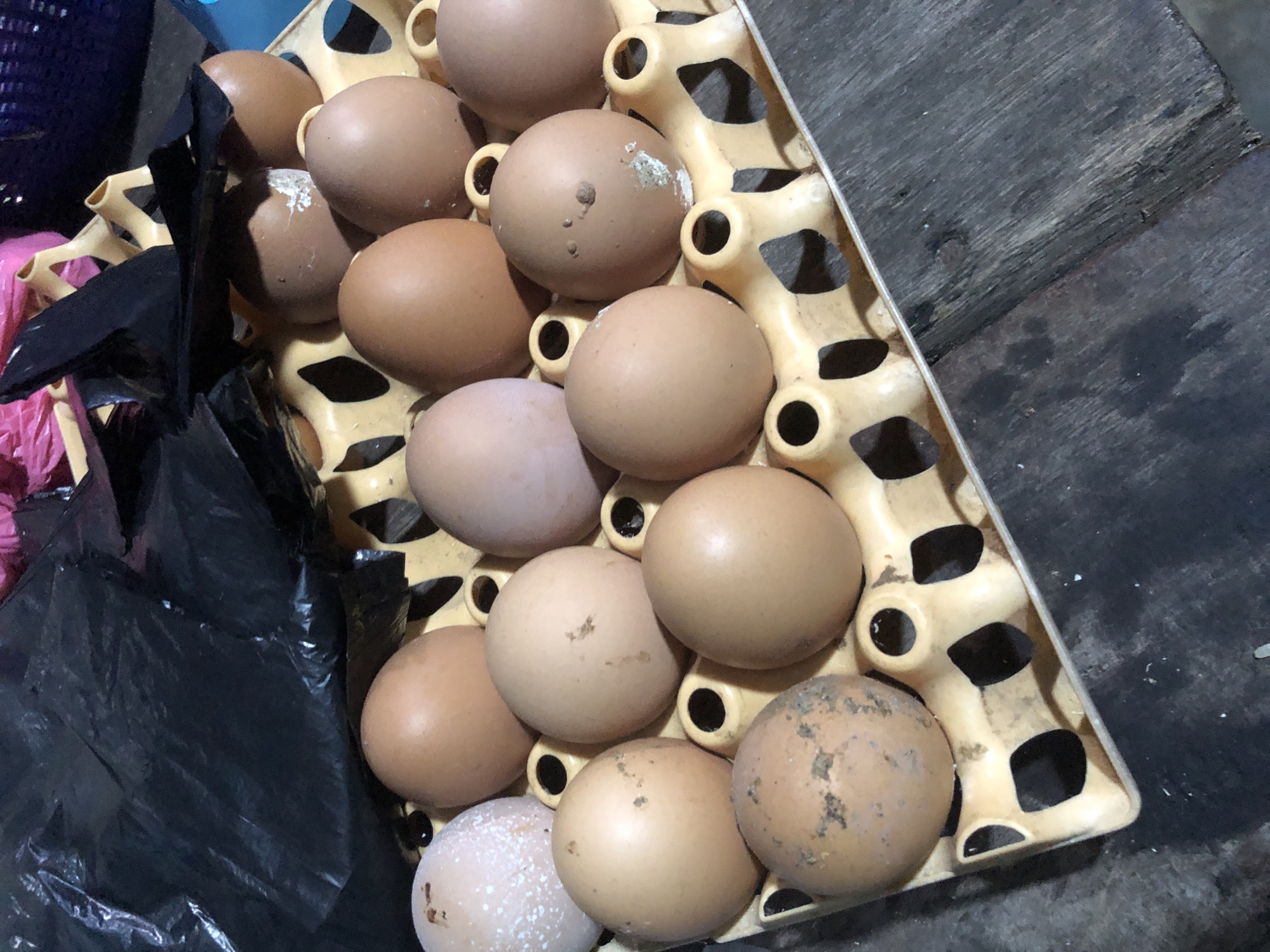 A dozen brown and white eggs are displayed in a yellow plastic egg carton. The carton is placed on a rough, weathered wooden surface, possibly a table or floor. The background shows a crumpled black plastic bag and hints of other colorful items, suggesting a market or storage area. The lighting is dim and appears to be artificial. The scene is likely located in Ado, Nigeria, given the provided context.