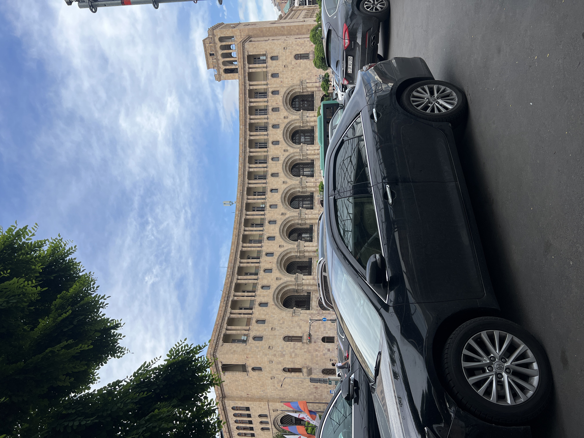 The image depicts a street scene in Yerevan, Armenia, with a focus on a large, light-brown, multi-story building featuring arched windows and architectural detailing. Several dark-colored cars are parked along the street, partially obscuring the building. The sky is blue with scattered white clouds. Green foliage is visible on the left side of the frame. Armenian flags are also visible.