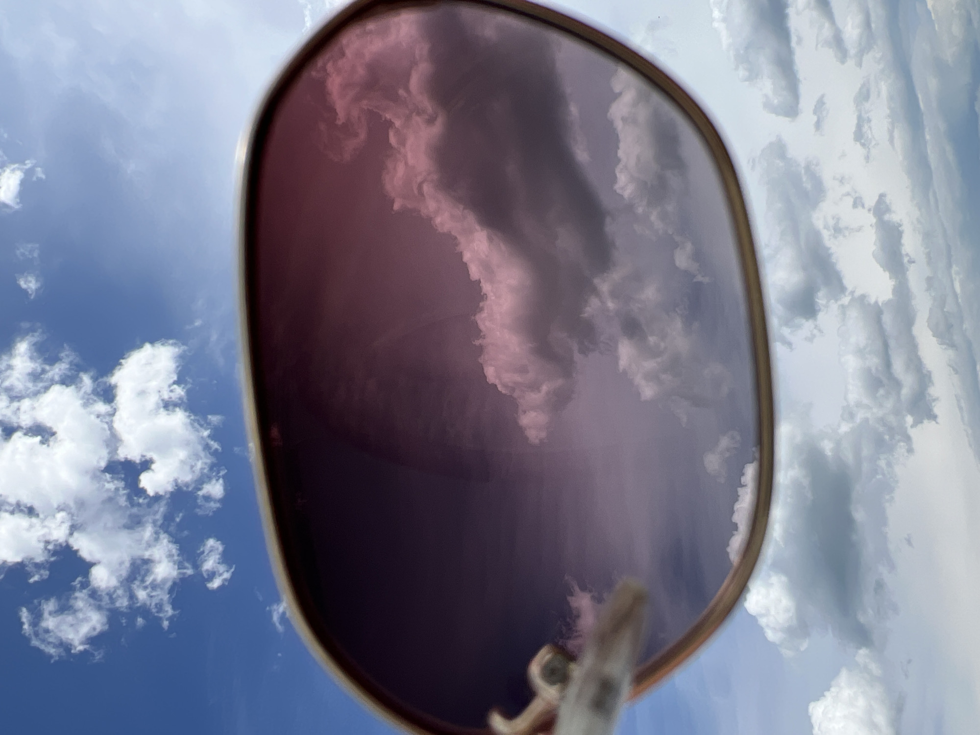 A pair of sunglasses with pink-tinted lenses is held up to the sky. The lenses reflect the sky, showing a mix of bright blue sky and fluffy white and pink-tinged clouds. The sunglasses frame appears to be gold-colored and is slightly out of focus. The background is a clear blue sky with scattered white clouds. There are no people or other objects clearly visible. The image does not provide any information to suggest a location in the city of Borychivka, Ukraine.
