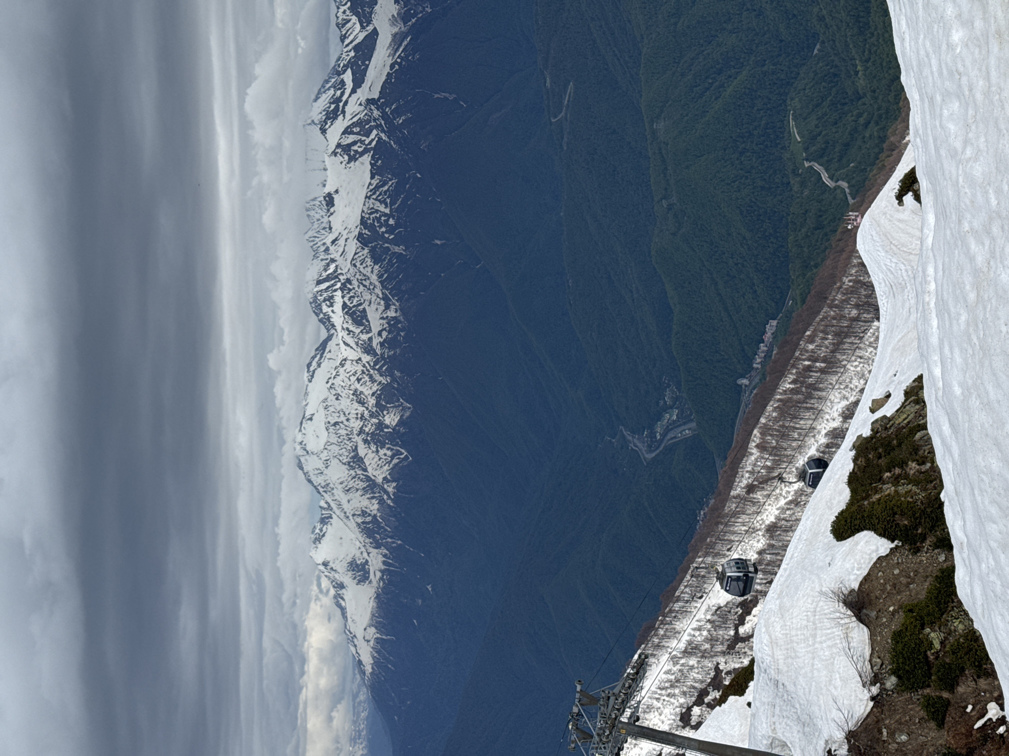 A vertical aerial shot taken from a high vantage point near Estosadok, Russia, shows a mountain landscape under a cloudy sky. The left side of the image features a mountain range with snow-capped peaks, which contrasts with a heavily forested valley. On the right, a cable car system with cars is visible descending a slope partially covered with snow and patches of greenery. The cable car tower is also visible.