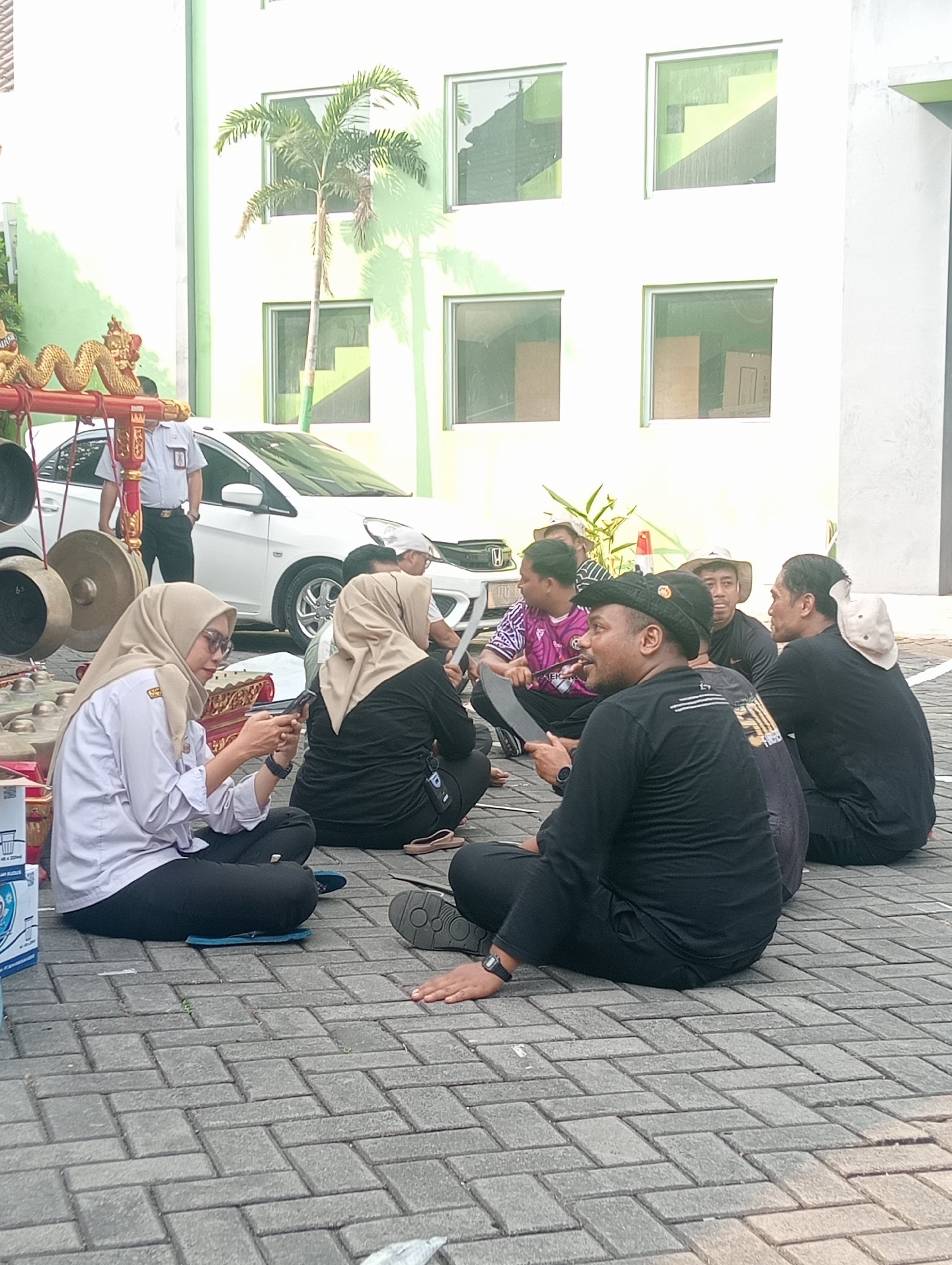 In Demak, Indonesia, a group of people, including men and women, are gathered outdoors on a paved area in front of a light green building with square windows. Several individuals are seated on the ground, some looking at their phones, while others are engaged in conversation. To the left, a traditional Gamelan musical instrument set is visible, with part of a white car and a person in uniform standing behind it. The overall atmosphere suggests a casual gathering, possibly related to a cultural event or performance in the city of Demak.