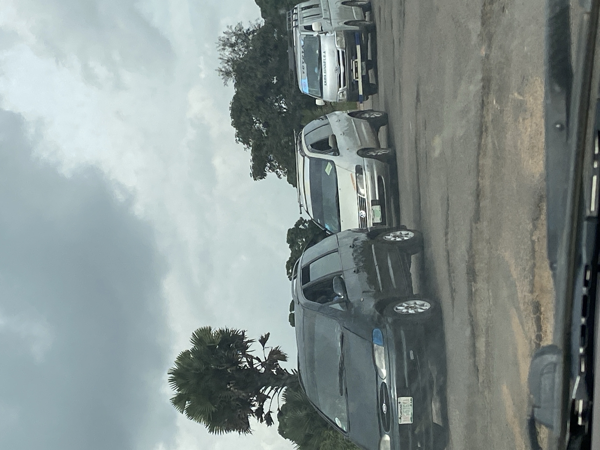 The image captures a traffic scene in Kuru, Nigeria. Three vehicles are visible on a damaged road, characterized by potholes. The leftmost vehicle is a dark-colored Ford minivan, its license plate reading "JAS-576KA". To its right is a silver Volkswagen minivan, and further to the right is a silver Toyota van, with "TOYOTA" text visible on its front. The sky is overcast with clouds, and trees, including a palm tree, are present in the background. The perspective suggests the photo was taken from inside another vehicle, looking forward through the windshield.