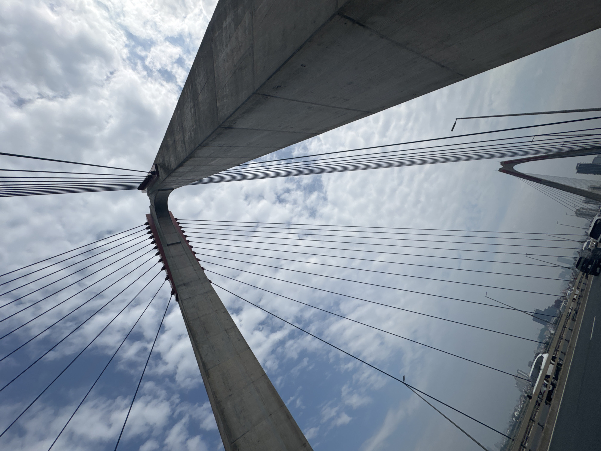 This is a low-angle, upward-facing, full shot of the Nhat Tan Bridge in Hanoi, Vietnam. The bridge is a cable-stayed bridge spanning the Red River. Two massive concrete pylons, painted red at the top, are visible, angling upwards and outwards, supporting numerous thin cables that extend downwards and outwards. The sky above is a mixture of blue with scattered white clouds. The roadway of the bridge is visible in the lower portion of the frame, with several vehicles, including cars and a bus, driving on it. In the distance, a hazy cityscape with tall buildings is discernible. The overall impression is one of modern infrastructure against a cloudy sky.