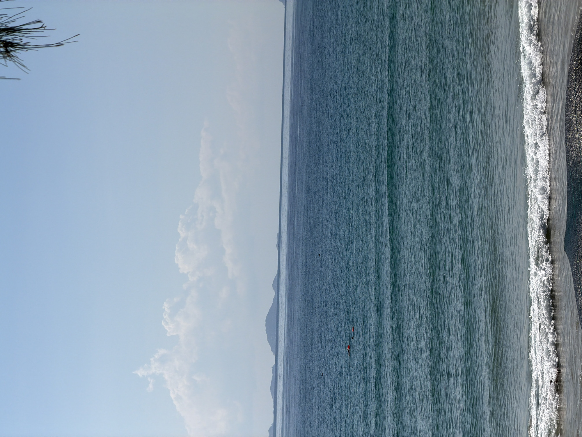 The image shows a beach in Cam Lam, Vietnam, taken during the daytime. The sky is blue with some white clouds. The sea is calm with gentle waves breaking on the shore. The beach appears to have dark sand. In the distance, there is a faint outline of land or islands. There are two red objects floating in the water. A tree branch is visible in the upper left corner.