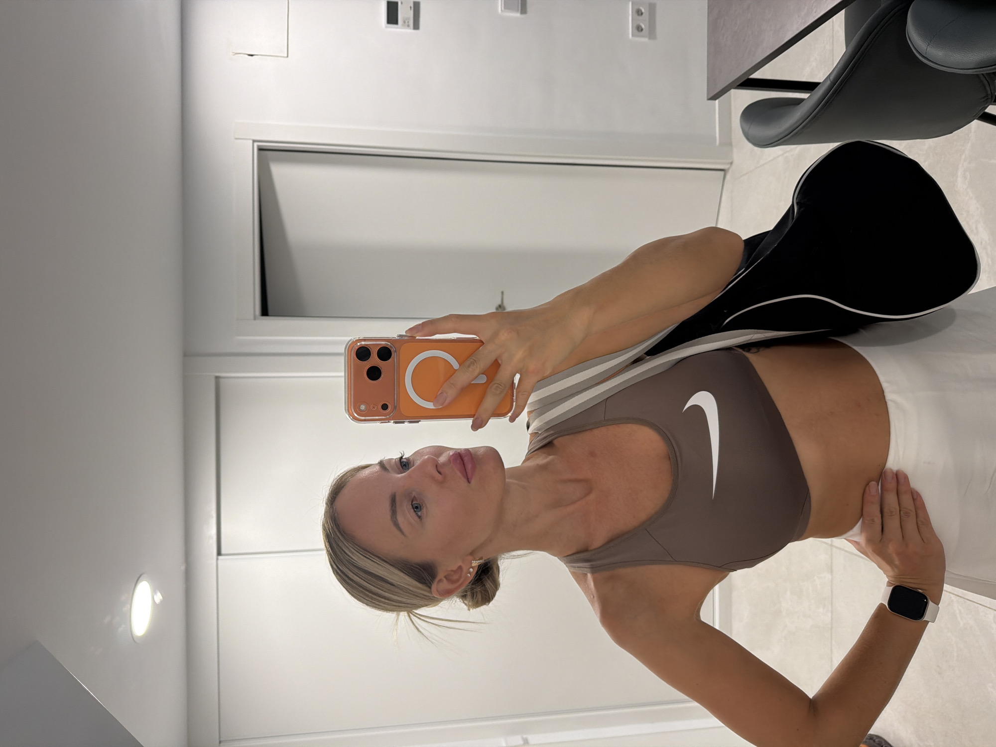 A fair-skinned woman with blonde hair pulled into a bun is taking a mirror selfie in what appears to be an indoor residential setting. She is wearing a brown Nike sports bra and a white skirt, with a black bag slung over her shoulder. She is holding an orange-cased iPhone in her right hand. The background shows white closet doors and a partially visible doorway. The floor is tiled. The scene does not contain any explicit indicators that it is located in Finestrat, Spain.