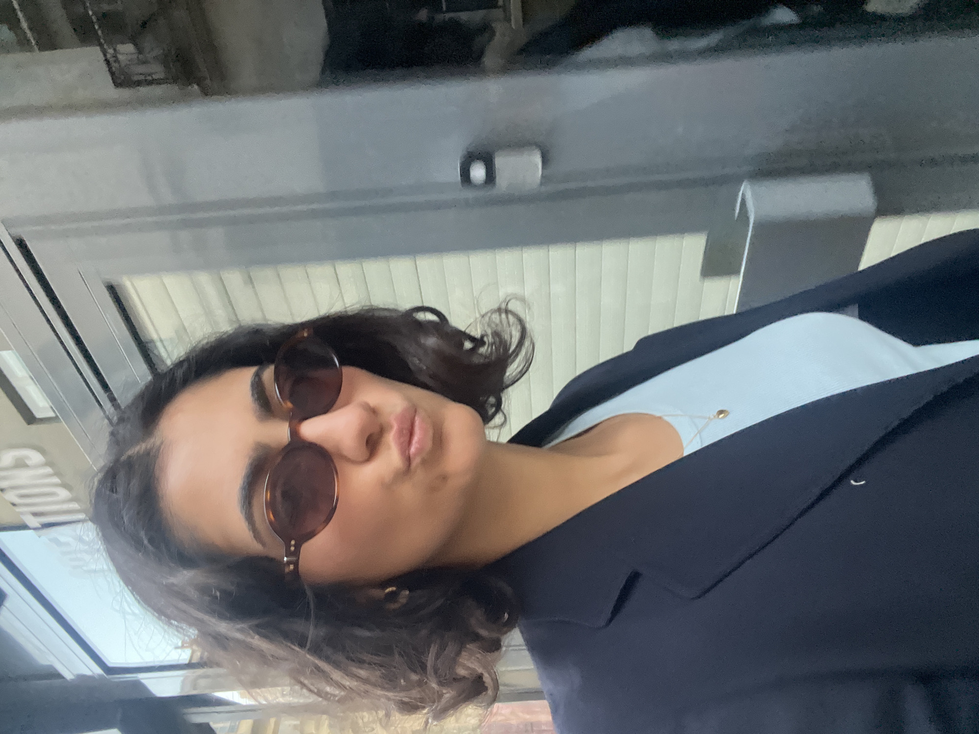 A woman with short, curly brown hair and wearing brown sunglasses is seen from the chest up. She is wearing a light blue top and a dark blue blazer. The background consists of a glass window or door with a sign and vertical blinds. Above the window or door is a gray, textured ceiling with a small black and white object. The reflection in the window suggests an urban environment, possibly in Paris, France.
