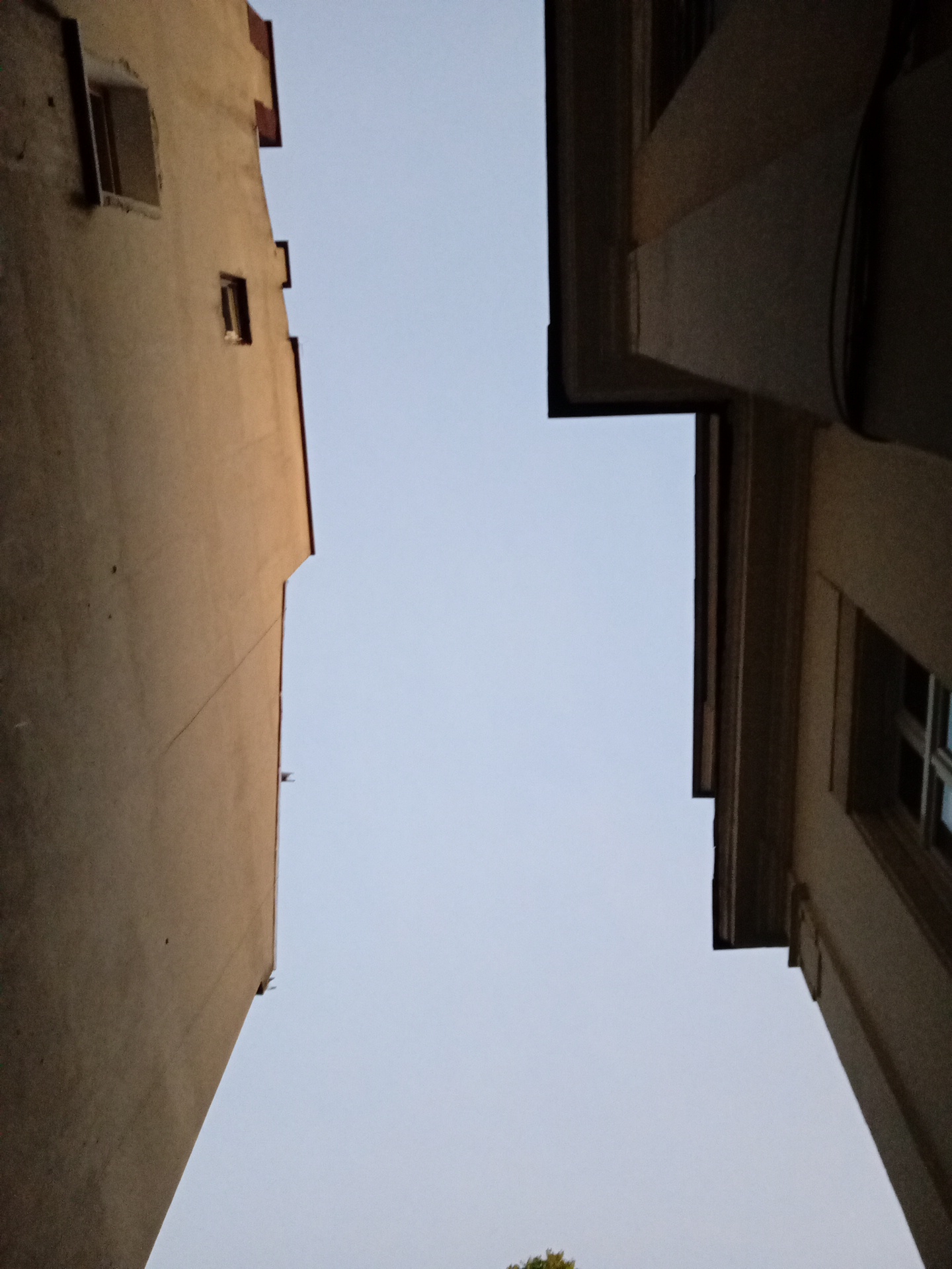 The image shows an upward view of two buildings facing each other, with a clear sky in between. The building on the left is a light brown structure with a visible window and architectural details on its upper edge. The building on the right is darker and features a window with visible panes. The scene is set in the city of Sofia, Bulgaria. There are no people or animals visible in the image.