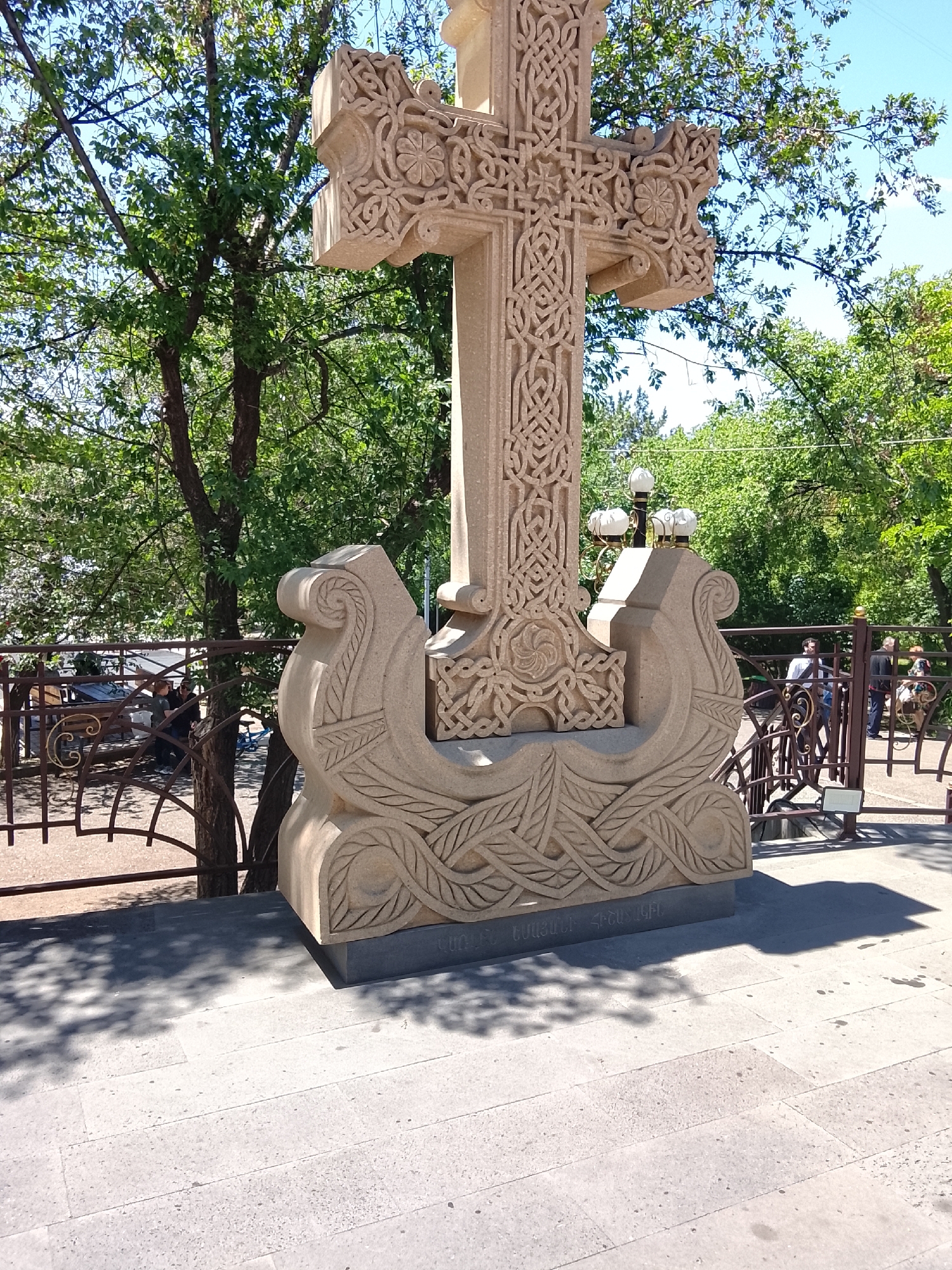 A large, intricately carved stone cross, known as a khachkar, stands prominently in an outdoor setting on a sunny day. The cross is made of light-colored stone and features elaborate patterns, including knotwork, geometric designs, and floral motifs, particularly noticeable on its arms and central shaft. Its base is also heavily carved with stylized wave-like or leafy designs, resembling the prow of a boat on each side, and sits on a darker stone plinth.
In the background, lush green trees with dense foliage provide shade, and a clear blue sky is visible above. A decorative metal fence or railing separates the foreground from the background activity. Several people are visible in the distance: some are seated at outdoor tables and chairs on the left, while others are standing or walking on the right, suggesting a public or semi-public area like a park or monastery grounds. A multi-branched lamppost with white, glowing globes is positioned behind the right side of the cross's base.
The ground is paved with light-colored square tiles, and distinct shadows of the cross and trees are cast upon it, indicating bright daylight. On the lower part of the khachkar's base, text carved in Armenian script is visible, which reads "ՍՈՒՐԲ ԷՋՄԻԱԾԻՆ ՏԱՃԱՐԻՆ", translating to "To Holy Etchmiadzin Cathedral". This inscription suggests the monument is dedicated to or located near the Mother See of Holy Etchmiadzin, Armenia.