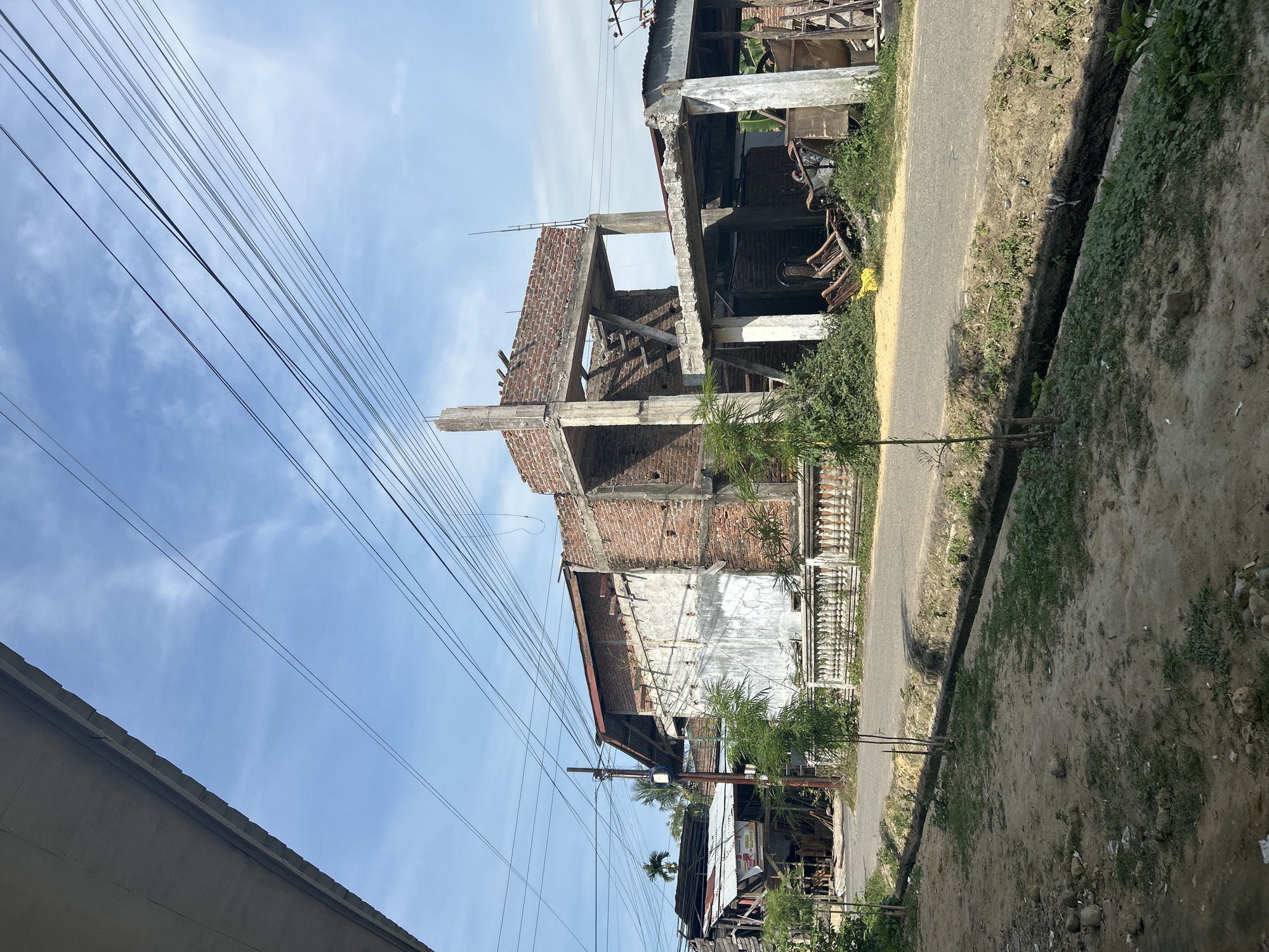 The image depicts a street scene in Lhokseumawe, Indonesia. On the left is a partially constructed or ruined building, mainly brick and concrete, with exposed supports and incomplete walls. Power lines are strung across the sky. The road runs along the right side of the building, bordered by a drainage ditch and dirt shoulder. Further down the road are other buildings, seemingly simple structures with a mix of materials. The sky is blue with some clouds.