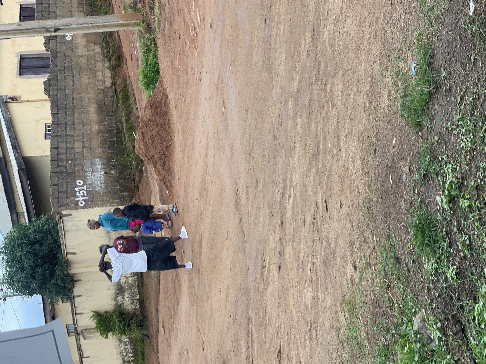 In Gwong, Nigeria, a group of five individuals, including three children and two adults, stand on a dirt road. The adult male in the foreground, wearing a white t-shirt and black shorts, has his hands on his head. To his right, an adult female in a teal hooded sweatshirt and an adult male in a maroon t-shirt and dark pants are also present. Two young boys, one in a blue shirt and the other in a black jacket, stand beside them. The background features a light yellow building with dark windows and a grey brick wall with white graffiti. A pile of reddish-brown dirt sits near the wall, and a utility pole is visible on the right side of the image. The ground is uneven and dusty, with sparse green vegetation growing in patches.