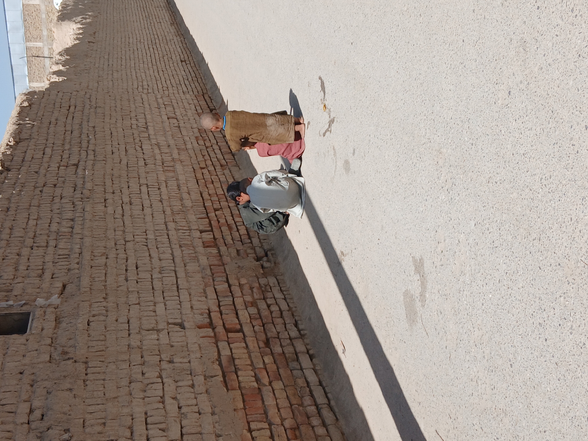 The image depicts an outdoor scene in Quetta City Tehsil, Pakistan, featuring two individuals next to a weathered brick wall adjacent to a paved area. One individual, older with a shaved head, is wearing a brown robe over what appears to be red clothing. The other, seemingly younger with dark hair, is wearing a gray robe and kneeling. The setting is stark, with sunlight casting shadows, emphasizing the textured surfaces of the wall and the ground. The brick wall shows signs of age and weathering. The paved area appears relatively clean and unobstructed.