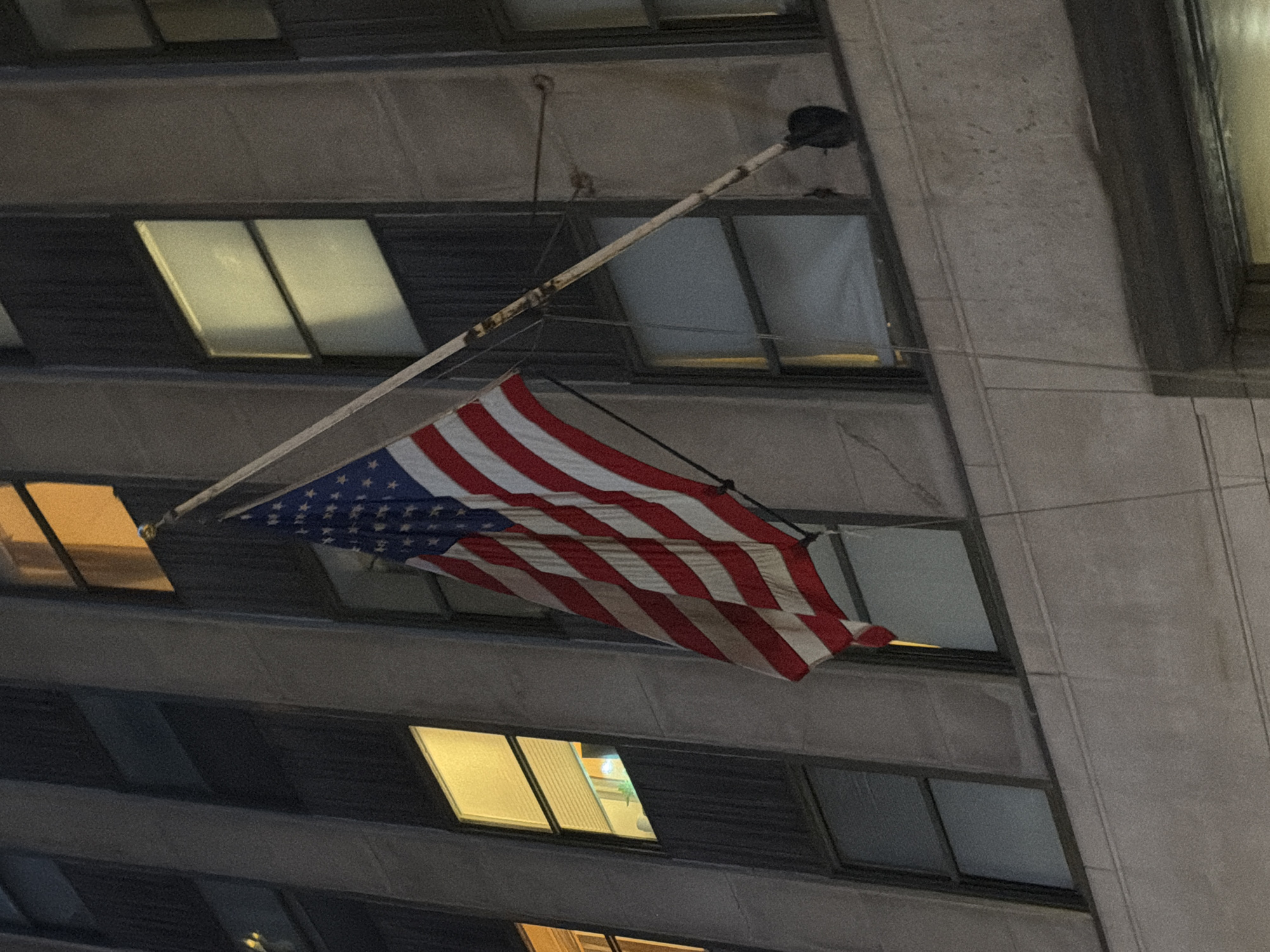 An American flag is attached to a flagpole mounted on the side of a building in New York City. The building appears to be an office building, with several windows visible, some of which are lit from within. The flag is partially furled and appears to be blowing in the wind. The image is taken at night, and the lighting suggests that the flag is illuminated by streetlights or internal building lights. The facade of the building is made of light-colored stone, and the windows are rectangular. The flag is red, white, and blue, with stars on a blue background. The flagpole is made of metal and appears to be old and weathered, with some rust visible.