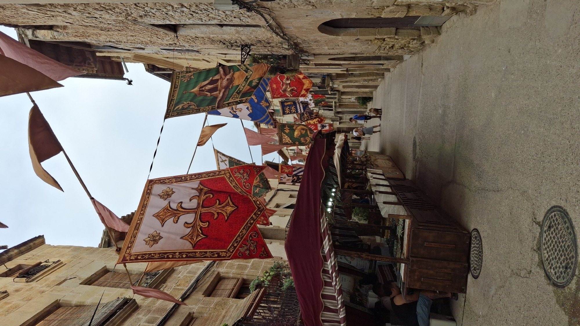 In La Fresneda, Spain, a narrow, cobblestone street is adorned with numerous colorful banners and flags hanging overhead, suggesting a festival or historical reenactment. The buildings flanking the street are constructed of stone, with balconies and shutters adding architectural detail. On the left side of the street, a market stall with a red awning displays various items for sale, with a person seated at one end. Further down the street, several individuals are walking, including a person with a dog. The sky is overcast, contributing to the diffused lighting of the scene. The overall atmosphere is one of a charming, historic town preparing for or hosting an event.