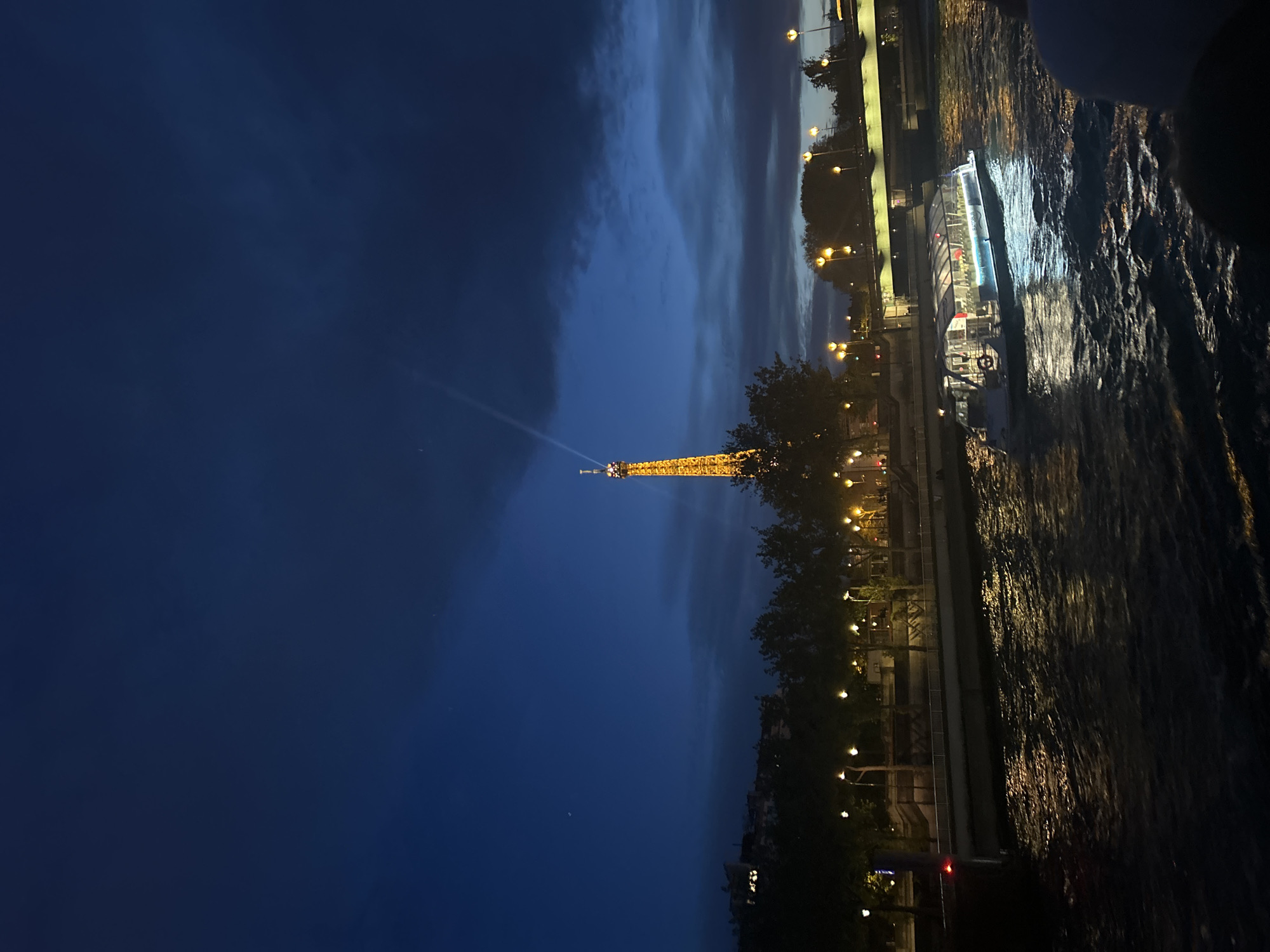 The image shows the Eiffel Tower at night, illuminated and emitting a beam of light. The tower is positioned along the Seine River. On the right of the image are the banks of the river and a boat. The sky is a mix of dark blue and grey clouds. There are trees and streetlights along the bank of the river. The image was likely taken from another boat on the river.