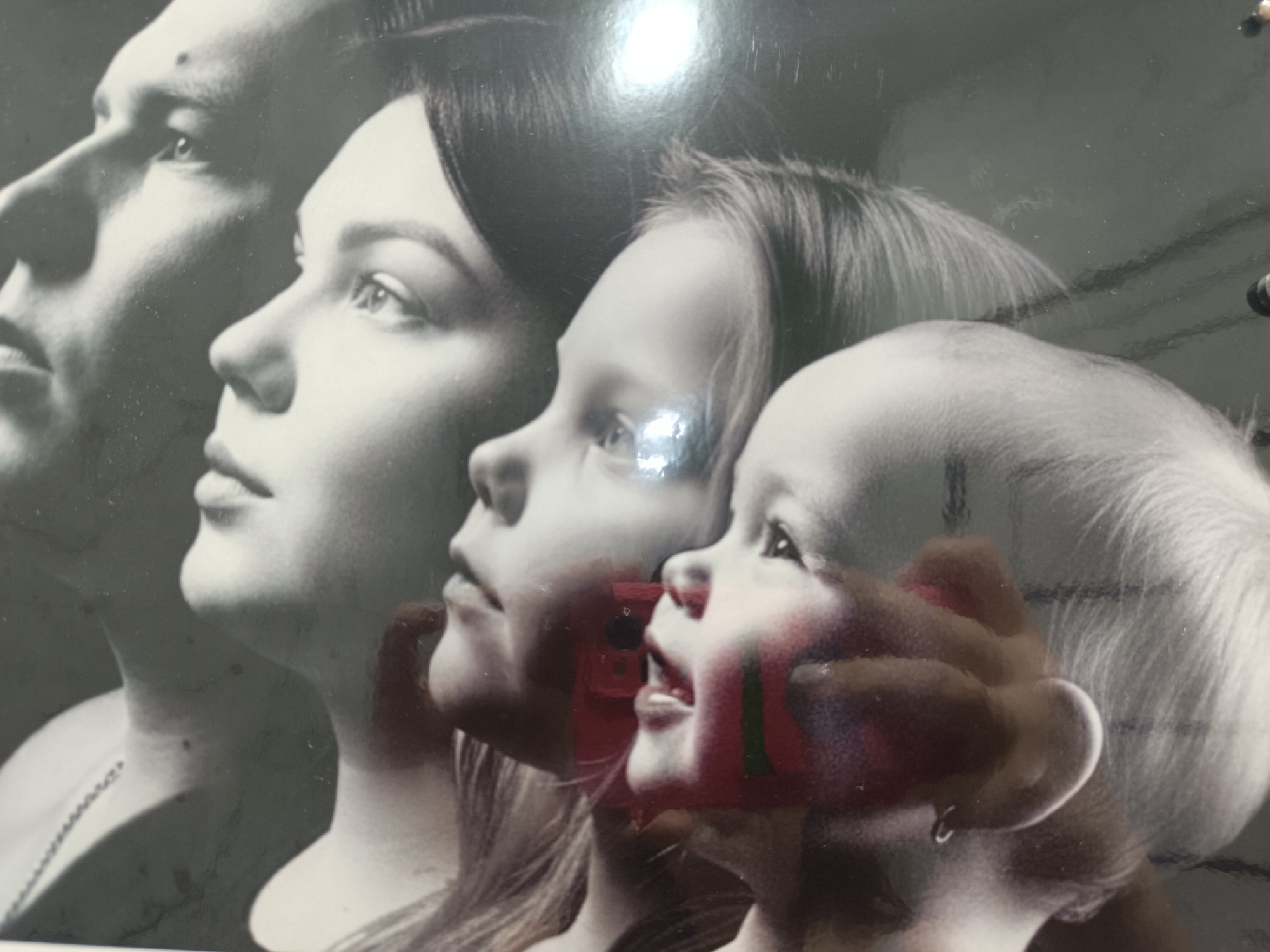 The image shows a black and white photograph of four individuals in profile, arranged from left to right. The subjects appear to be a family: a man, a woman, a young girl, and a baby. They are positioned closely together, with each face partially obscuring the next. The photograph has reflections across the surface. The presence of the city of Chaikovsky, Russia is unknown.