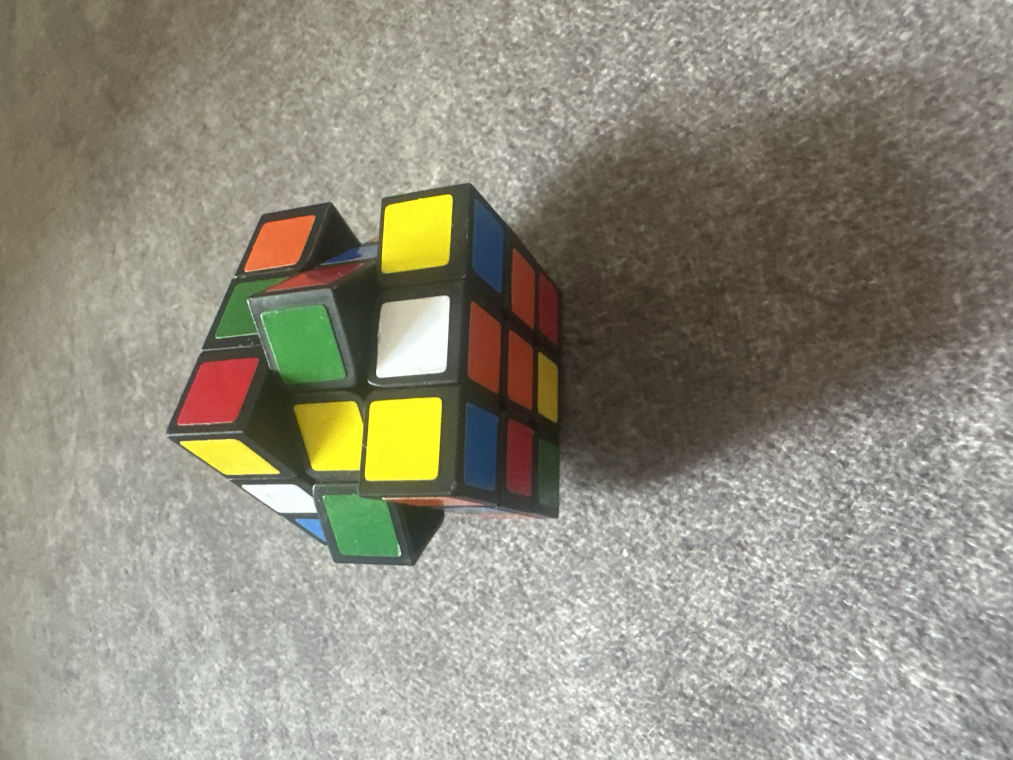 A partially scrambled Rubik's cube rests on a grey, textured surface, likely carpet or a similar textile. The cube displays various colors including red, yellow, green, blue, white, and orange. The lighting suggests an indoor environment, casting a subtle shadow to the right of the cube. No people are present in the image, and there are no discernible landmarks or indications of a specific city or location.