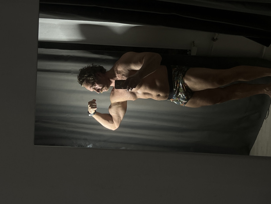 A shirtless man with curly hair flexes in a mirror, taking a selfie. He wears patterned briefs and a wristwatch. The mirror reflects a shadowy room with a gray wall behind him and a dark fabric backdrop above. The lighting is dim, highlighting his physique.