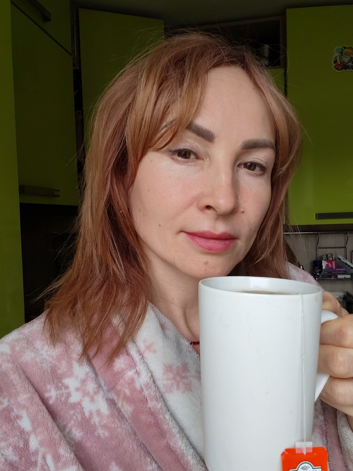 A woman is holding a white mug with a teabag in it. She is wearing a pink and white patterned robe. The background shows green kitchen cabinets. The location is possibly Chaikovsky, Russia.