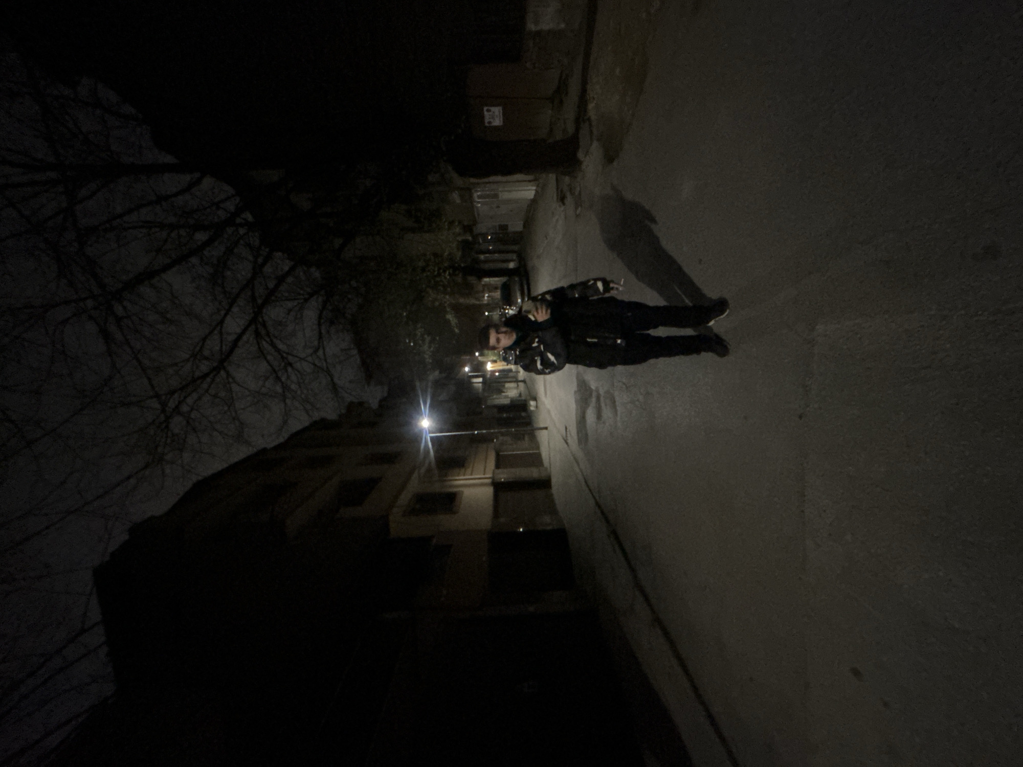 At night in Plovdiv, Bulgaria, a person stands on a paved street illuminated by distant streetlights and nearby buildings. The individual is wearing a dark jacket, dark pants, and dark shoes. The background includes bare trees on the left and buildings of varying heights. The scene is dark, with low visibility. The perspective is skewed as if the picture was taken at an angle.