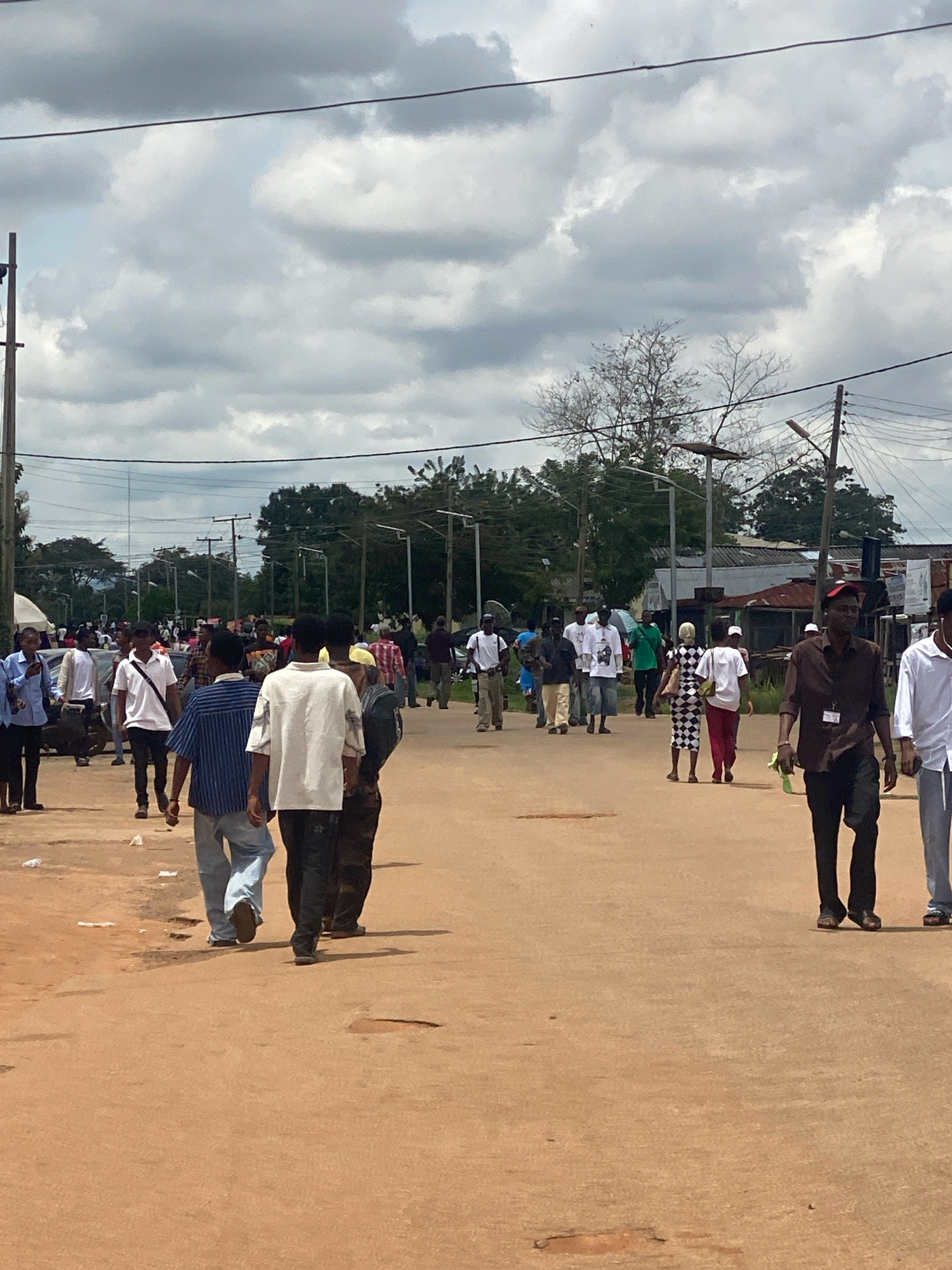 The image depicts a street scene in Iworoko, Nigeria, with a significant number of people walking along a dusty, unpaved road. The sky is overcast with heavy clouds. In the background, there are trees, utility poles with overhead wires, and some buildings, including what appears to be a market area with stalls and roofs. Several cars are visible, parked or driving slowly. The people are dressed in casual attire, and a few are carrying bags. The overall atmosphere suggests a bustling day, possibly a market day or a community gathering.