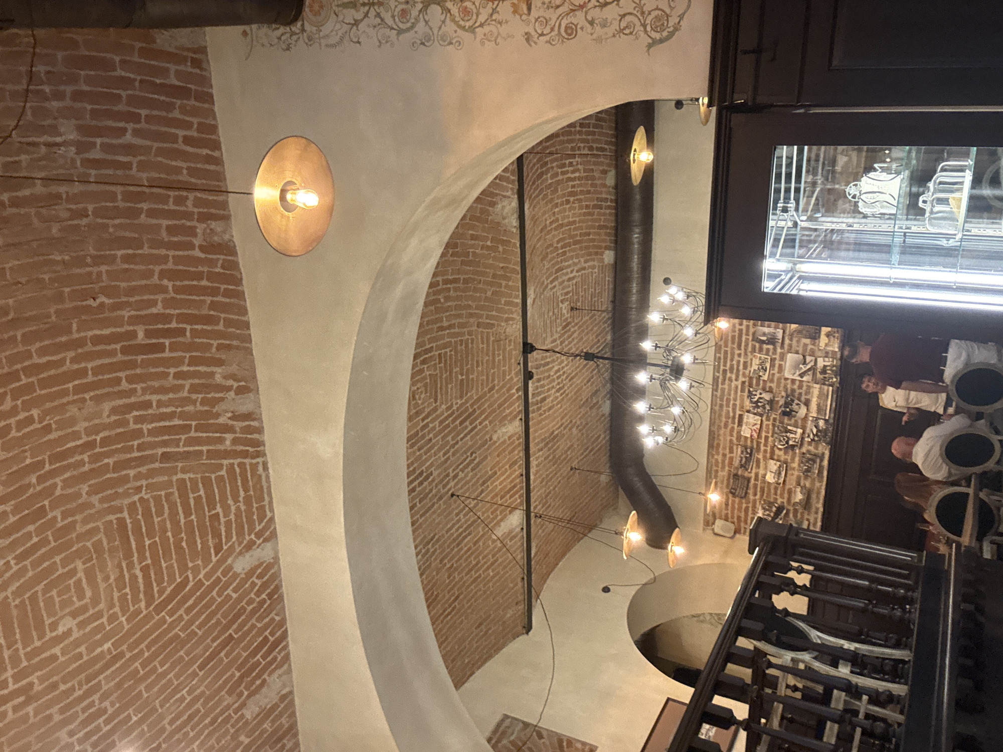 This media file captures an indoor scene, likely a restaurant or bar, with a distinct architectural style. The primary subjects are the interior elements of the space. Exposed brickwork forms curved walls and vaulted ceilings, creating a warm and rustic ambiance. Several light fixtures are visible, including a large, modern chandelier with multiple bulbs, and smaller, industrial-style pendant lights with exposed bulbs and copper-colored shades. The lighting suggests it is likely evening or nighttime, as the interior is illuminated by artificial light.
In the background, a staircase with dark, ornate railings leads upwards. A glass display cabinet can be seen, showcasing various items, possibly related to the establishment's theme or products. Several people are present in the background, standing near a doorway and a wall adorned with framed pictures or documents. Their attire and the general atmosphere suggest a social gathering or dining experience. The overall impression is of a well-designed, perhaps historical, venue. There is no visible text within the frame that can be readily deciphered. The location context provided indicates Chernivtsi, Ukraine, which might further inform the architectural style and decor if the viewer has knowledge of local design trends. The weather is not determinable from the image.