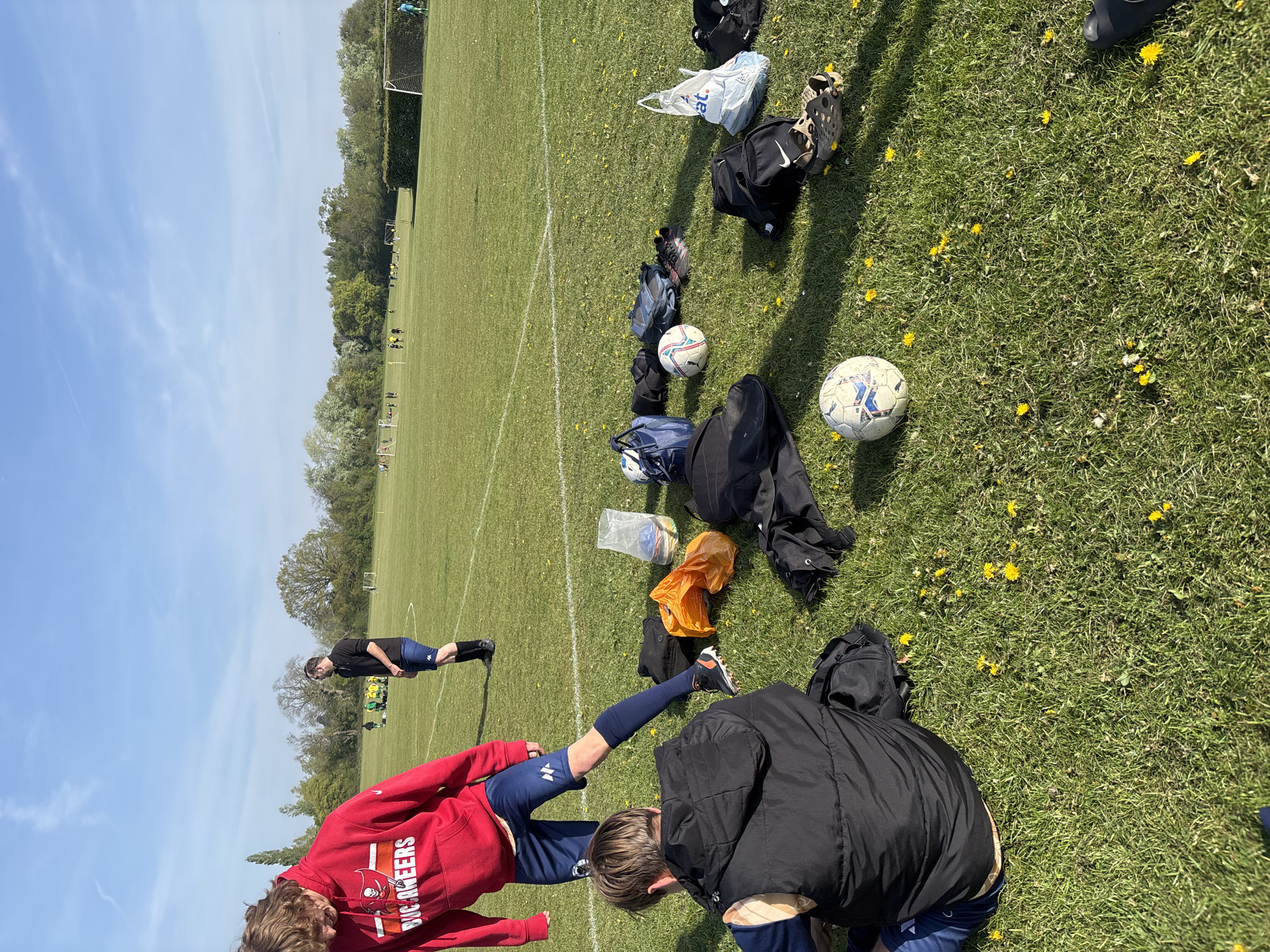 The image shows a grassy field with yellow dandelions, likely a sports field. There are soccer balls and bags scattered on the grass. In the foreground, two people are present: one wearing a red hoodie with "BUCCANEERS" printed on the back is stretching their leg, while another person in a black jacket is bending down. In the background, another person is stretching. Beyond them, a soccer game appears to be in progress in the distance. A white line marks the field and there are trees in the distance. The sky is blue with light clouds. The location is likely St Albans, United Kingdom.
