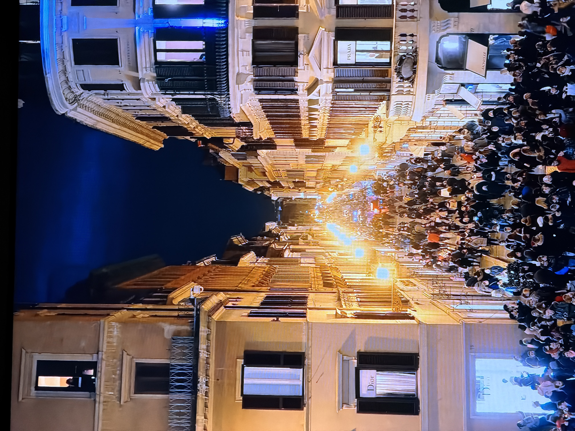 The image depicts a crowded street scene at night, illuminated by streetlights and lights from the buildings. Tall, multi-story buildings line both sides of a narrow street. The architecture appears European, with ornate detailing and shuttered windows. Several storefronts are visible, including Dior and Prada, indicating a high-end shopping district. A large number of people are walking down the street, some in small groups, others individually. The sky is dark blue. The scene suggests a vibrant urban environment, possibly a popular tourist destination. The city of Chirchik, Uzbekistan is not depicted in this image.