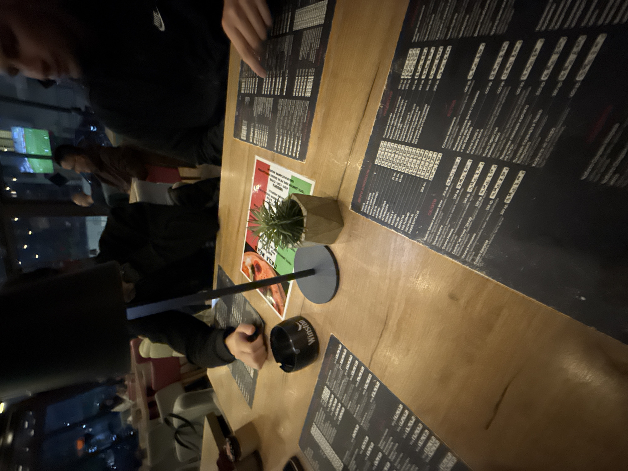 The image shows a table at what appears to be a restaurant in Pernik, Bulgaria. Multiple people are seated around the wooden table, with menus from a restaurant titled "PIZZA MENU" (Пица Меню) visible. There is an ashtray from the brand "Winston" on the table. A table decoration with a small plant is present. Some of the menu text is in Bulgarian. The room is indoors and dimly lit. There are lights and a TV screen visible in the background, possibly showing a sports game. People are looking at menus.