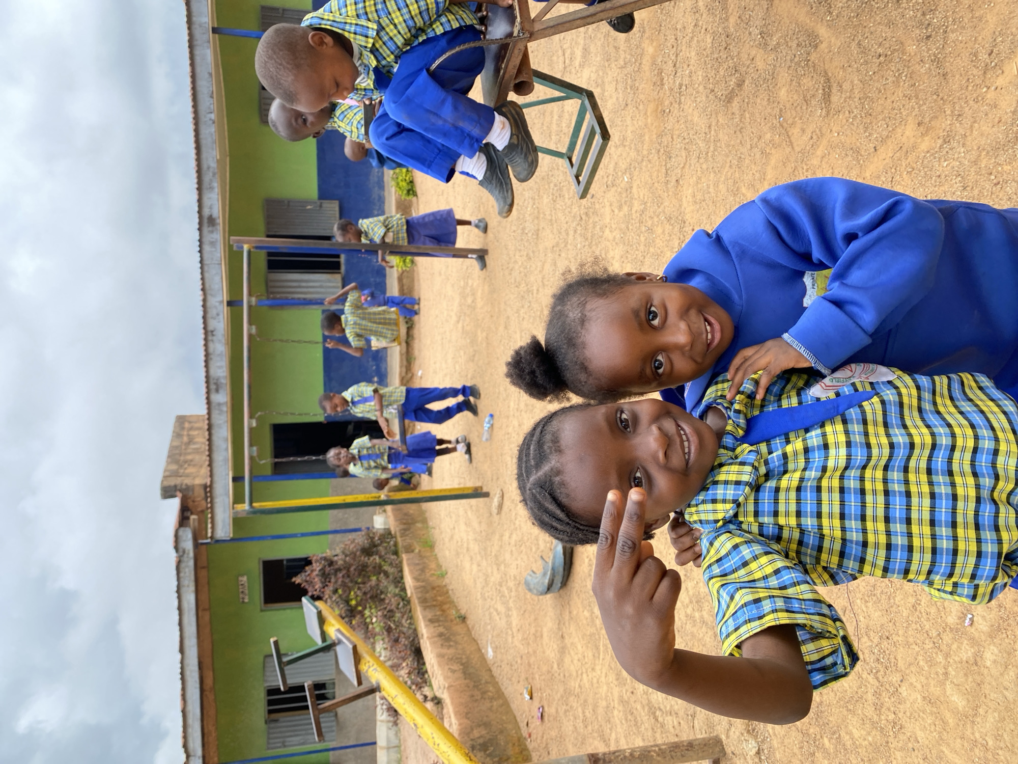 The image, taken in Gura Top, Nigeria, shows two schoolgirls posing in the foreground with other children in the background on a playground. One girl wears a blue and yellow plaid shirt and makes a peace sign with her fingers; the other wears a blue jacket. In the background, other children in similar school uniforms play on swings and a merry-go-round in front of a low, green and blue building. The playground has a sandy ground surface.