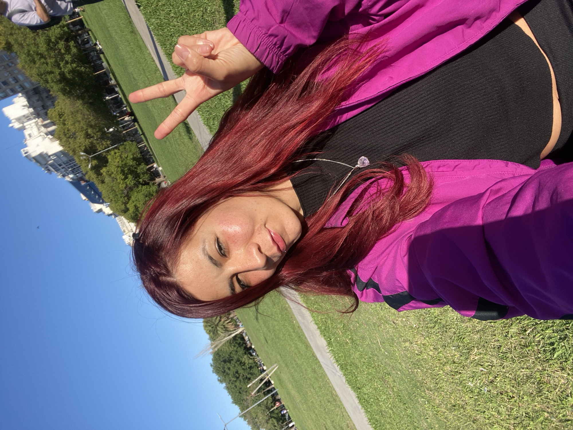 The image is a selfie taken in a park. The subject is a young woman with red hair and wearing a pink jacket over a black top. She is making a peace sign with her hand. In the background, there is a green field with a path. Beyond the field, there are trees and buildings. The buildings and trees suggest the location is in a city park. Specifically, the buildings in the background, the layout of the park, and the green spaces suggest this image was taken in Parque Tres de Febrero in Buenos Aires, Argentina.