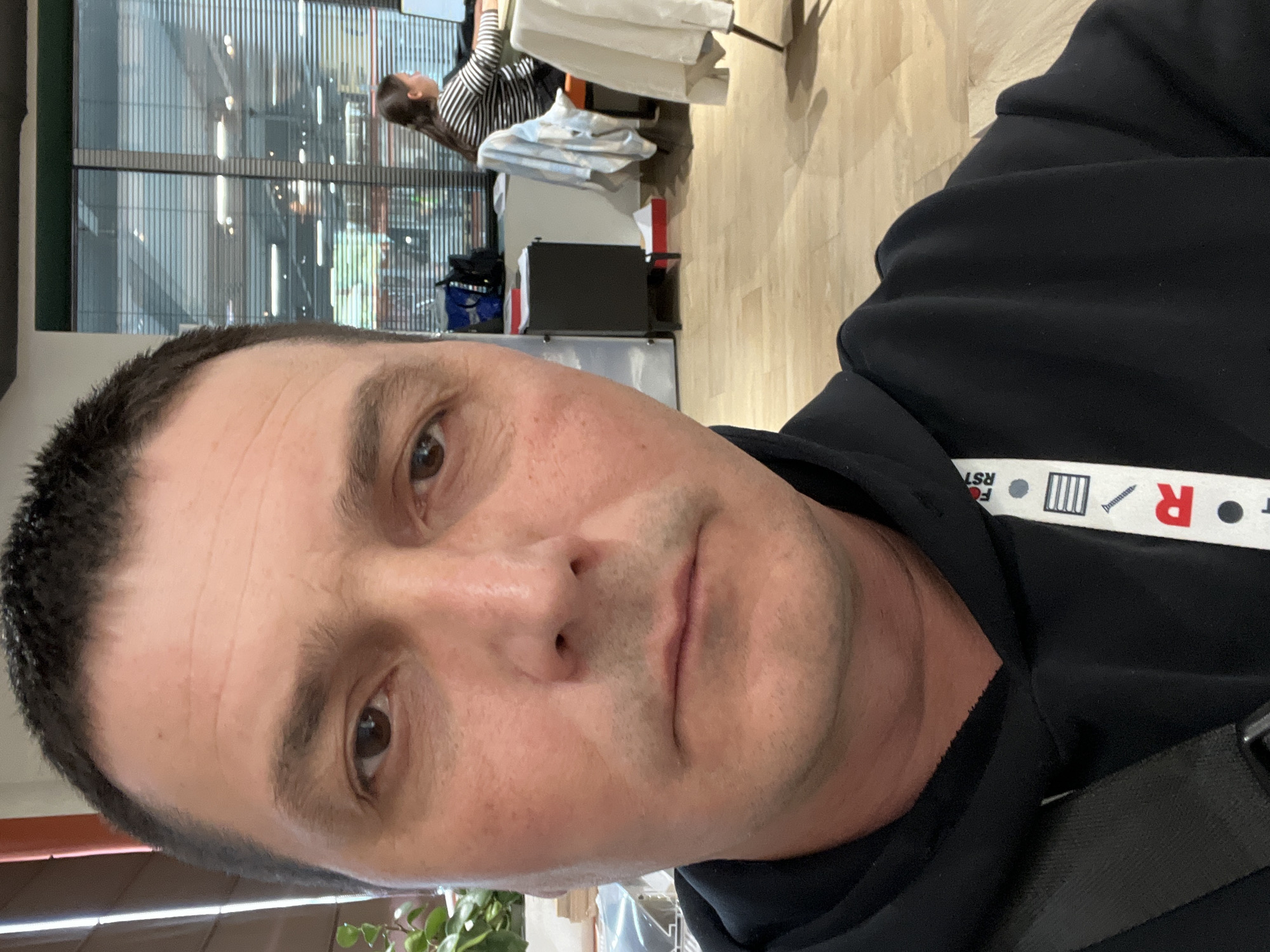 A man is depicted in a close-up selfie-style shot, angled to the right, looking directly at the camera. He has short, dark hair, brown eyes, and a neutral expression. He is wearing a black hoodie. A white lanyard with text and symbols hangs around his neck; visible elements include "RST", partially "FOR", a symbol resembling three vertical lines within a rectangle, what appears to be a screw, and a large red "R". A black strap, possibly from a bag, is visible over his right shoulder.
In the background, an indoor setting is visible, suggesting a modern office, co-working space, or public building. Another person with dark hair and a black-and-white striped long-sleeved top is seen in the mid-ground, leaning over a white chair or object. This person appears to be engaged in an activity at a table or desk. The background features large windows or glass panels with vertical elements, letting in natural light, indicating it is daytime. The floor is made of light-colored wood or laminate. Scattered objects like a blue bag and part of a plant are also discernible in the background.