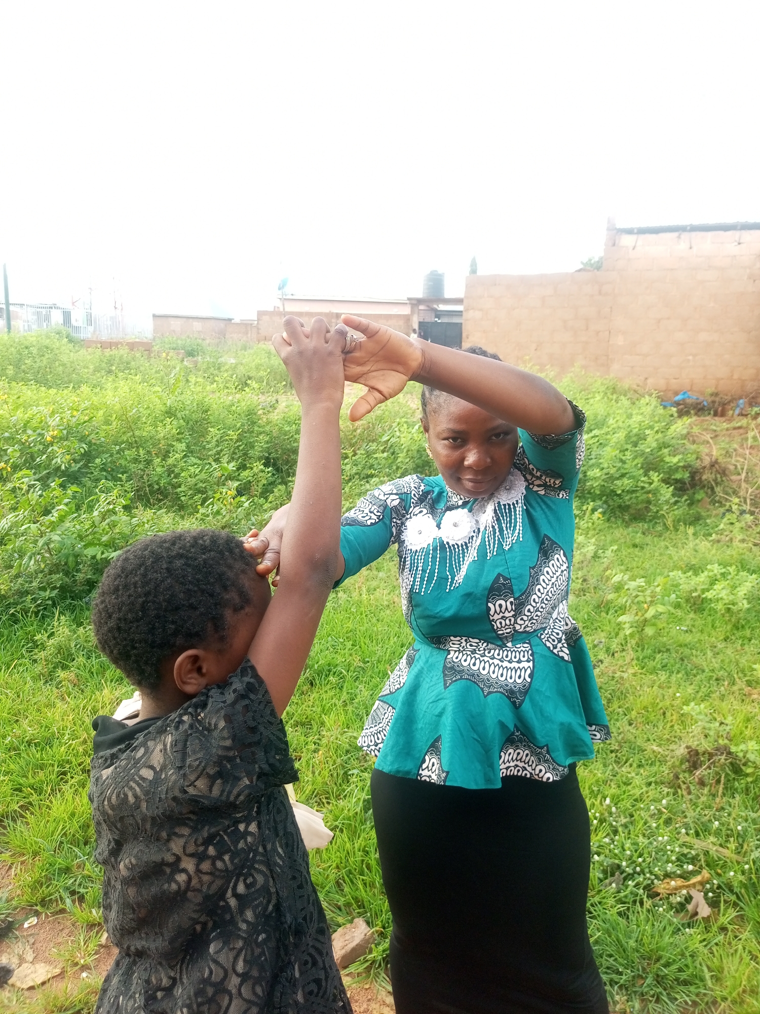 In a rural or semi-urban outdoor setting in Tempure, Nigeria, two individuals, likely an adult woman and a child, are engaged in an interaction. The adult woman, positioned in the center-right of the frame, wears a teal and white patterned peplum top and a black skirt. She is facing the camera and has her left arm raised, with her hand near her head. To her left and slightly in front, the child, seen from the back, is wearing a black lace dress. The child's head is turned away from the camera, and their arms are also raised, with their hands reaching towards the adult's arms. The background consists of green vegetation, some scattered bricks, and buildings in the distance, suggesting a residential or undeveloped area. The sky is overcast and bright, indicating a daytime scene.