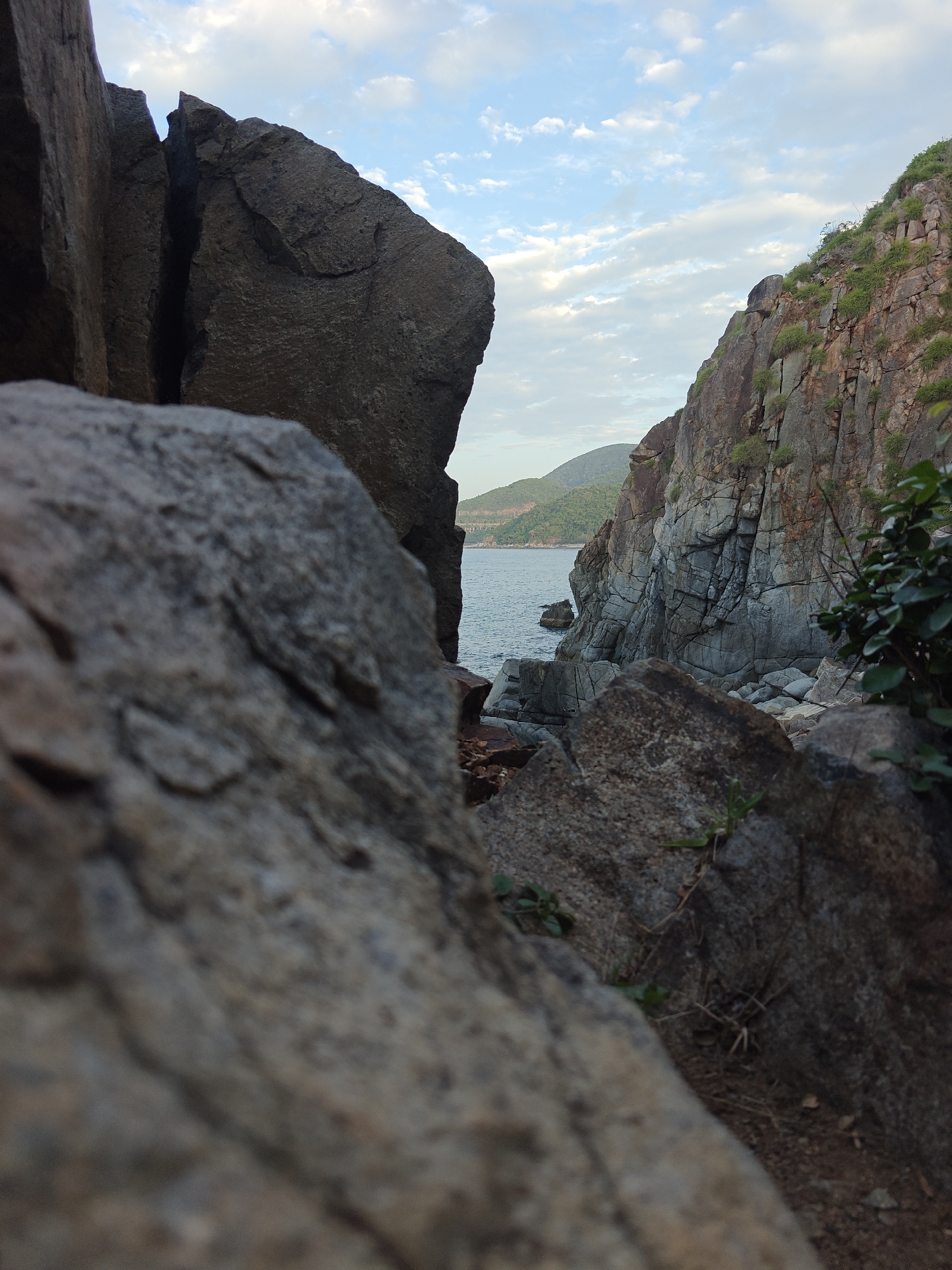 This is a low-angle, close-up shot of a rocky coastline near Vĩnh Lương, Vietnam. The foreground is dominated by large, textured rocks, one of which is sharply in focus and takes up the left half of the frame. Beyond these rocks, a body of water, likely the sea, is visible, meeting a distant, tree-covered hill and a rugged, grey cliff face. The sky above is a light blue with scattered white clouds. The overall impression is one of natural, rugged beauty.