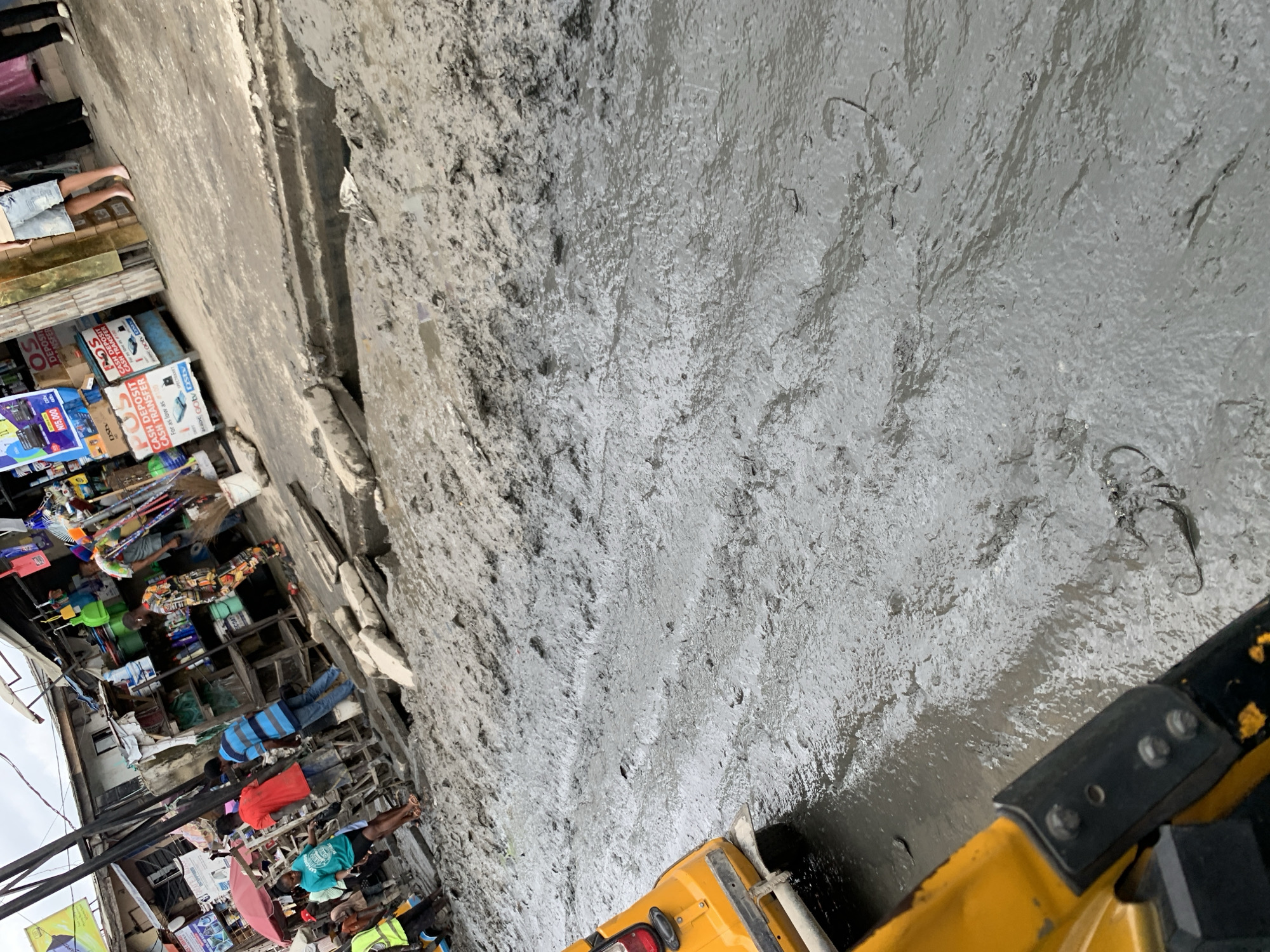 This image depicts a muddy street scene in Ado, Nigeria, likely after heavy rainfall. A yellow motorized rickshaw is partially visible on the left side of the frame, suggesting it is navigating the difficult terrain. The street is covered in thick, grey mud, with visible tracks from vehicles and possibly pedestrians. In the background, market stalls are set up along the side of the road, with various goods displayed. Several people are present, some appearing to be vendors or shoppers. One individual in a red shirt and another in a blue and striped shirt are walking through the mud. Other individuals are seen in the background near the market stalls. The overall environment appears to be an outdoor market with limited infrastructure, as indicated by the unpaved and muddy road. The sky is overcast, suggesting the possibility of continued rainfall.