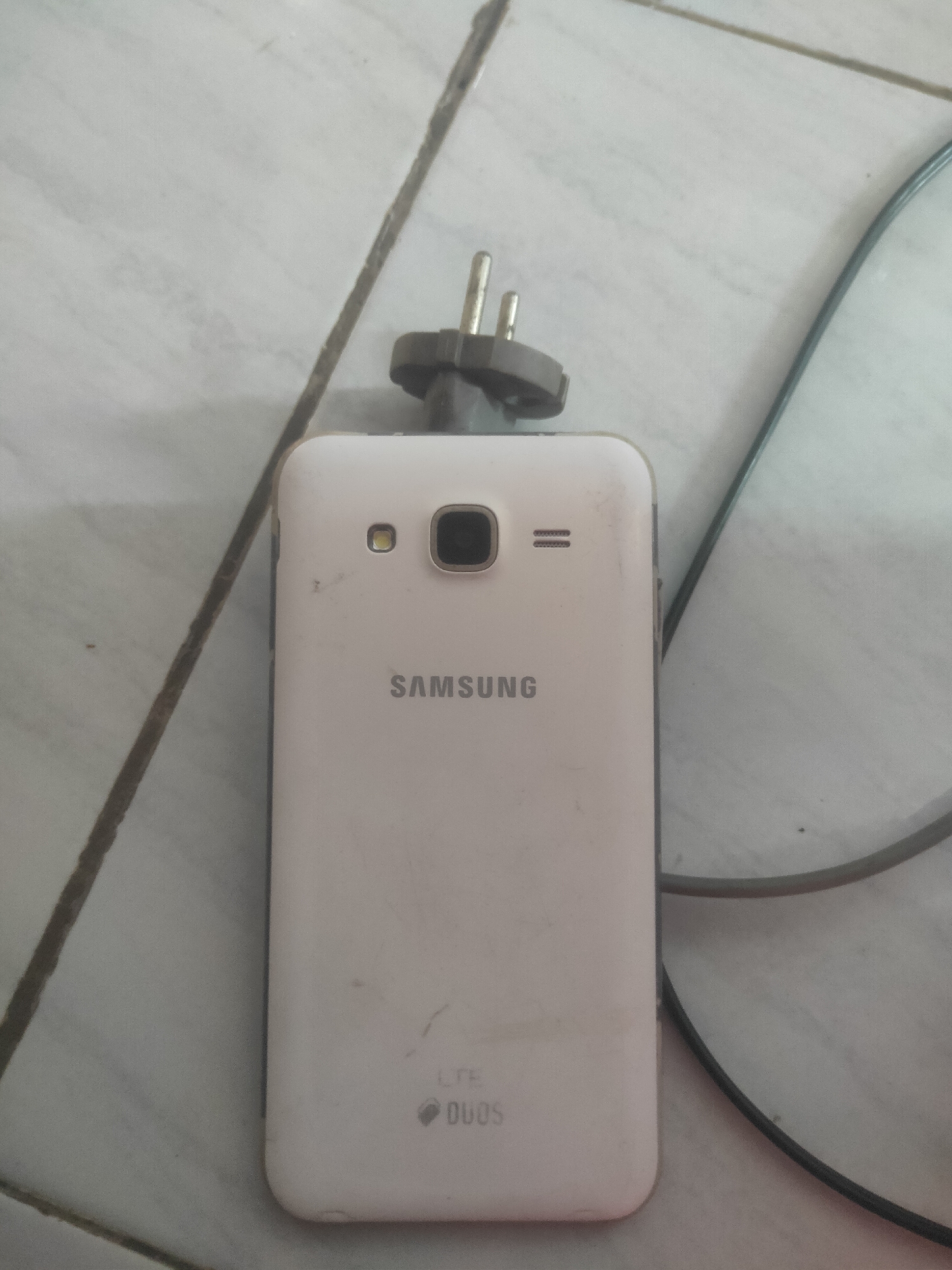 A white Samsung smartphone is placed on a light-colored tiled surface. The phone is facing upwards, revealing its back panel. A power adapter is plugged into the top of the phone, with its prongs extending upwards. A black cable is connected to the power adapter and extends out of the frame. The background consists of light-colored tiles with darker grout lines. The image is likely taken in Antirogo, Indonesia.